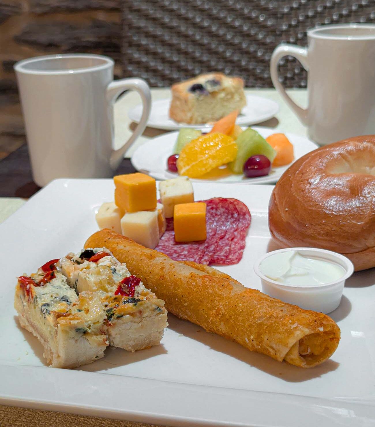A close-up view of a continental breakfast plate from the Lancaster Arts Hotel, showcasing their complimentary morning offerings. The white plate in the foreground features an array of breakfast items including a savory quiche or egg dish, a crispy breadstick or krisproll, cubes of cheese (both yellow cheddar and white varieties), slices of salami, and what appears to be a portion of cream cheese or spread with a small container of dipping sauce. In the background is another plate with fresh fruit including orange slices, kiwi, and cranberries, alongside what looks like a blueberry muffin or pastry. Two white coffee mugs complete the presentation. The setup demonstrates the hotel's attention to quality and variety mentioned in the travel guide, where the continental breakfast is described as including made-to-order pancakes, Mediterranean quiche, fresh fruit, yogurt, and scones.