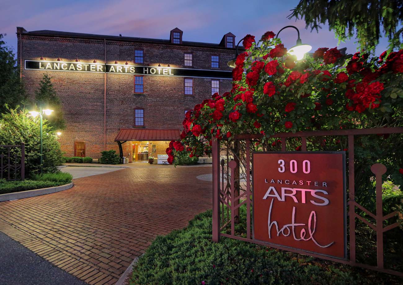 Evening exterior view of the Lancaster Arts Hotel, housed in a beautifully restored 19th-century brick tobacco warehouse. The building is softly illuminated with its name displayed in white lights across the facade. In the foreground, vibrant red roses bloom on trellises beside the hotel's entrance sign, a nod to Lancaster's nickname as the "Red Rose City." The brick driveway leads to the entrance, which is warmly lit and welcoming. Landscaped gardens with shrubs and small trees frame the property. This twilight image captures the hotel's blend of historic industrial architecture and artistic sensibility, showcasing how Lancaster has repurposed its historic structures for contemporary use. The prominent "300" address marker and "Arts Hotel" signage in stylized lettering reflects the property's identity as not just accommodation but a living gallery with over 260 original pieces of art by Pennsylvania artists.