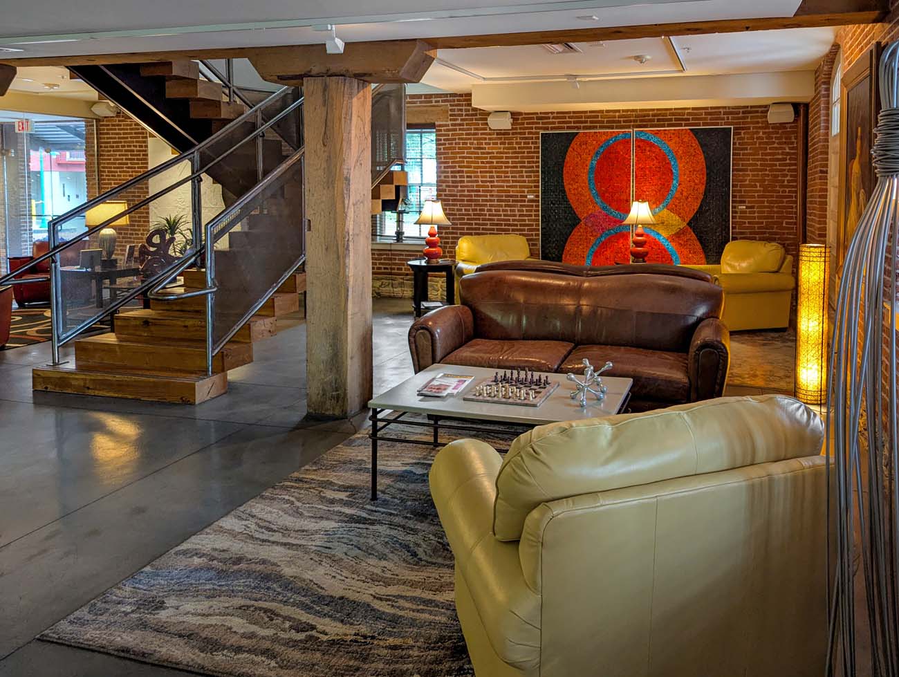 Interior view of the Lancaster Arts Hotel lobby, showcasing its artistic character and adaptive reuse of the historic tobacco warehouse. The space features original exposed brick walls, wooden support beams, and concrete floors that maintain the building's industrial heritage. The seating area includes a brown leather sofa and yellow leather armchairs arranged around a coffee table with a chess set, creating a comfortable gathering space. A striking colorful abstract artwork dominates the brick wall in the background, demonstrating the hotel's commitment to showcasing Pennsylvania artists throughout the property. A modern staircase with glass and metal railings leads to upper levels, while warm lighting from contemporary fixtures creates an inviting atmosphere. This image illustrates why the Lancaster Arts Hotel is described in the travel guide as "a mix of history, charm, and creativity" where the building itself is part of the artistic experience.