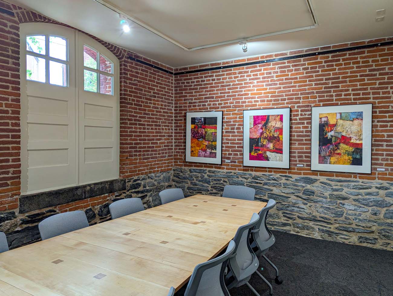 A meeting room or private dining space at the Lancaster Arts Hotel that perfectly blends historic architecture with contemporary art and design. The room features dramatic exposed brick walls and a lower section of original stone foundation, showcasing the building's 19th-century industrial origins. Three vibrant abstract paintings in bright reds, yellows, and blacks are displayed against the brick wall, exemplifying the hotel's collection of over 260 original pieces by Pennsylvania artists. A wooden conference table with six modern gray chairs occupies the center of the space, suitable for small meetings or private dining. Natural light enters through a white-framed door with multiple window panes. The space demonstrates how the Lancaster Arts Hotel preserves historical elements while functioning as a living gallery, creating a unique environment that celebrates both Lancaster's architectural heritage and its thriving contemporary arts scene.