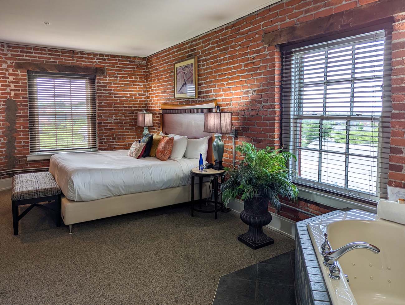Interior view of a spacious bedroom of the Presidential Suite at the Lancaster Arts Hotel, highlighting the original exposed brick walls that reflect the building's history as a 19th-century tobacco warehouse. The room features a king-sized bed with white linens and colorful accent pillows, wooden headboard, and stylish bedside lamps with blue-green ceramic bases. Large windows with horizontal blinds allow natural light while offering views of the surroundings. A potted fern adds a touch of greenery, and a corner of a jetted tub is visible, likely part of the Presidential Suite mentioned in the blog post. The preserved historic elements like the brick walls are seamlessly combined with modern comforts, exemplifying the hotel's commitment to honoring Lancaster's industrial past while providing contemporary luxury.