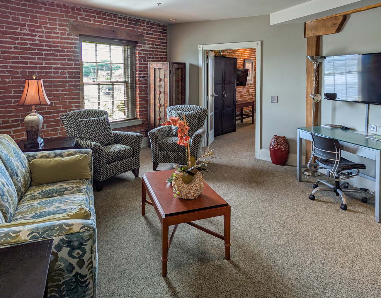 A well-appointed living area in a suite at the Lancaster Arts Hotel, showcasing the property's blend of historic character and modern comfort. The space features a patterned sofa with green accent pillows, two chevron-patterned armchairs, and a wooden coffee table. One wall displays the building's original exposed brick with a window overlooking downtown Lancaster, while other walls are painted in a soft sage green. Rustic wooden support beams are visible in the doorway and ceiling, preserving the building's original tobacco warehouse structure. A desk with an office chair sits against the right wall below a mounted TV, creating a functional workspace. The room is decorated with tasteful lamps and floral accents, reflecting the hotel's artistic character mentioned in the blog post as a "living gallery with over 260 original pieces by Pennsylvania artists."