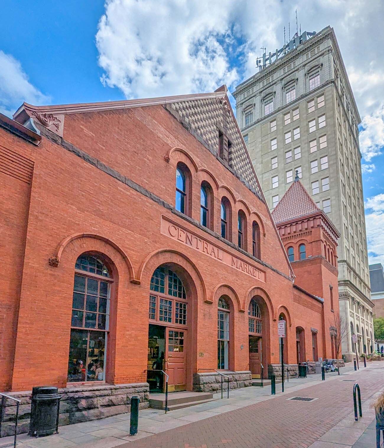 Exterior view of the iconic Lancaster Central Market building with its distinctive red brick Romanesque Revival architecture. The words "CENTRAL MARKET" are carved into the facade above a series of arched windows and entrances. The historic market building features decorative brickwork, stone foundation, and a prominent gabled roof with patterned detail. In the background, the modern Griest Building tower rises against a bright blue sky with white clouds, creating a striking contrast between Lancaster's historic and contemporary architecture. This image showcases the market that has been operating since 1730, making it America's oldest continuously running farmers market as mentioned in the travel guide. The pedestrian-friendly brick paving in front highlights the walkable nature of downtown Lancaster.