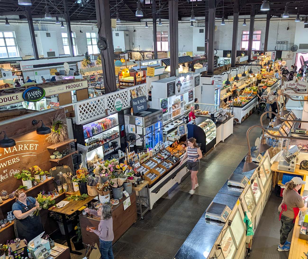 Overhead view of the vibrant interior of Lancaster Central Market, displaying the bustling marketplace with its numerous vendor stalls arranged in orderly rows. The historic industrial building features high ceilings with exposed structural elements and pendant lighting. Visible vendor stalls include "Barr's Farm Fresh Produce" and several food and flower stands. Shoppers can be seen browsing the market, with colorful displays of fresh fruits, vegetables, baked goods, and specialty foods. The market's layout demonstrates its function as a community gathering place where over 65 vendors, many representing multi-generational family businesses, offer everything from farm-fresh ingredients to prepared foods. This image captures the authentic experience described in the blog post as one of Lancaster's must-visit attractions, showing why the author recommends bringing a cooler bag to take home treasures from this historic marketplace.