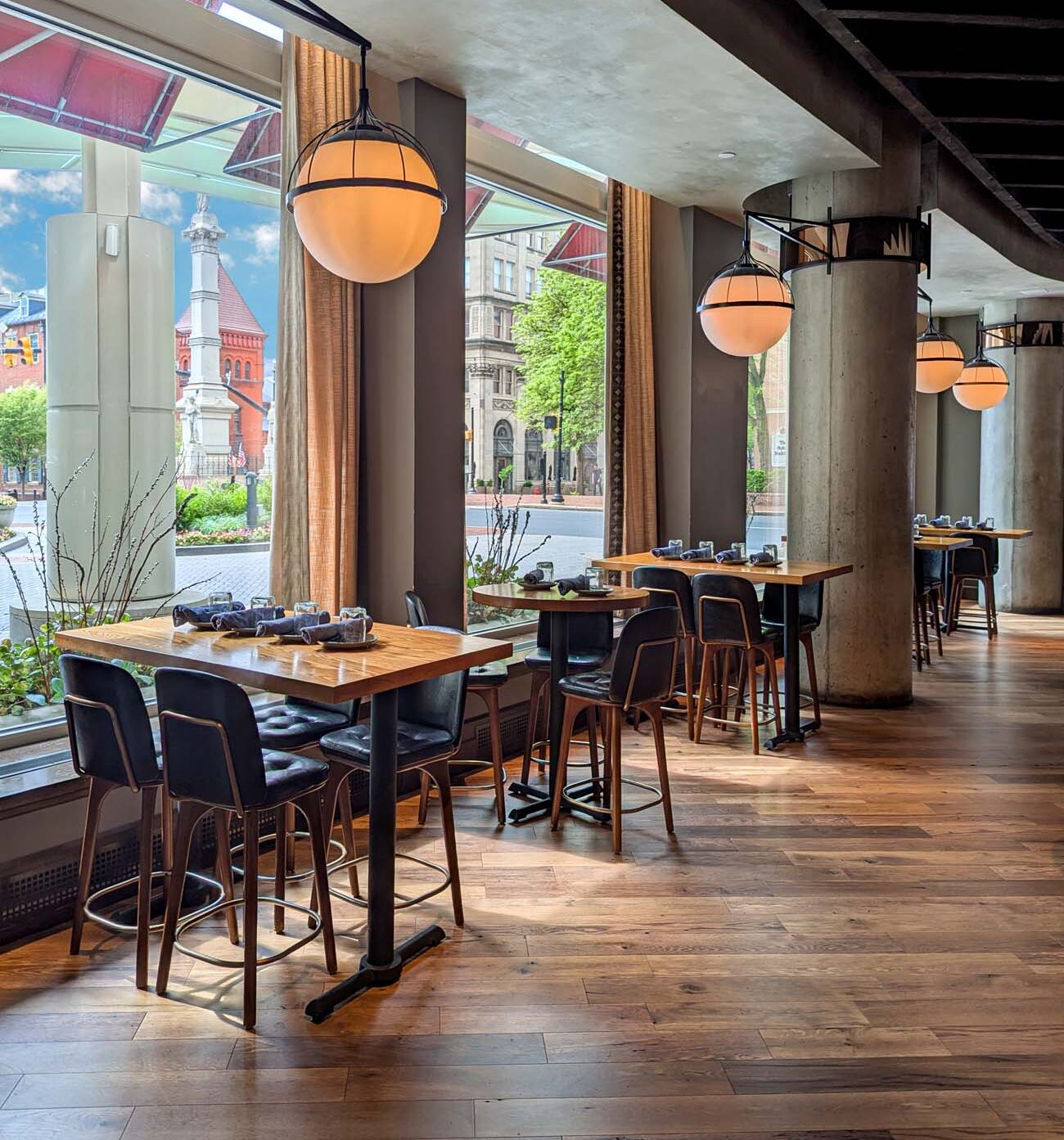 Interior view of the Plough Restaurant inside the Lancaster Marriott at Penn Square, showcasing its modern design and prime location. High-top wooden tables with leather bar chairs line floor-to-ceiling windows that overlook Penn Square and the historic Soldiers and Sailors Monument. Warm globe pendant lights hang from the ceiling, creating an inviting atmosphere against the restaurant's concrete columns and wood flooring. The contemporary space features a mix of industrial and warm elements, with curtains softening the windows and carefully placed greenery adding life to the space. Through the windows, downtown Lancaster's historic architecture is visible in the daylight. This image captures the "high ceilings, warm wood floors, and floor-to-ceiling windows offering perfect views of historic downtown" mentioned in the blog post's description of Plough, illustrating why it makes an excellent starting point for exploring the city's culinary scene.