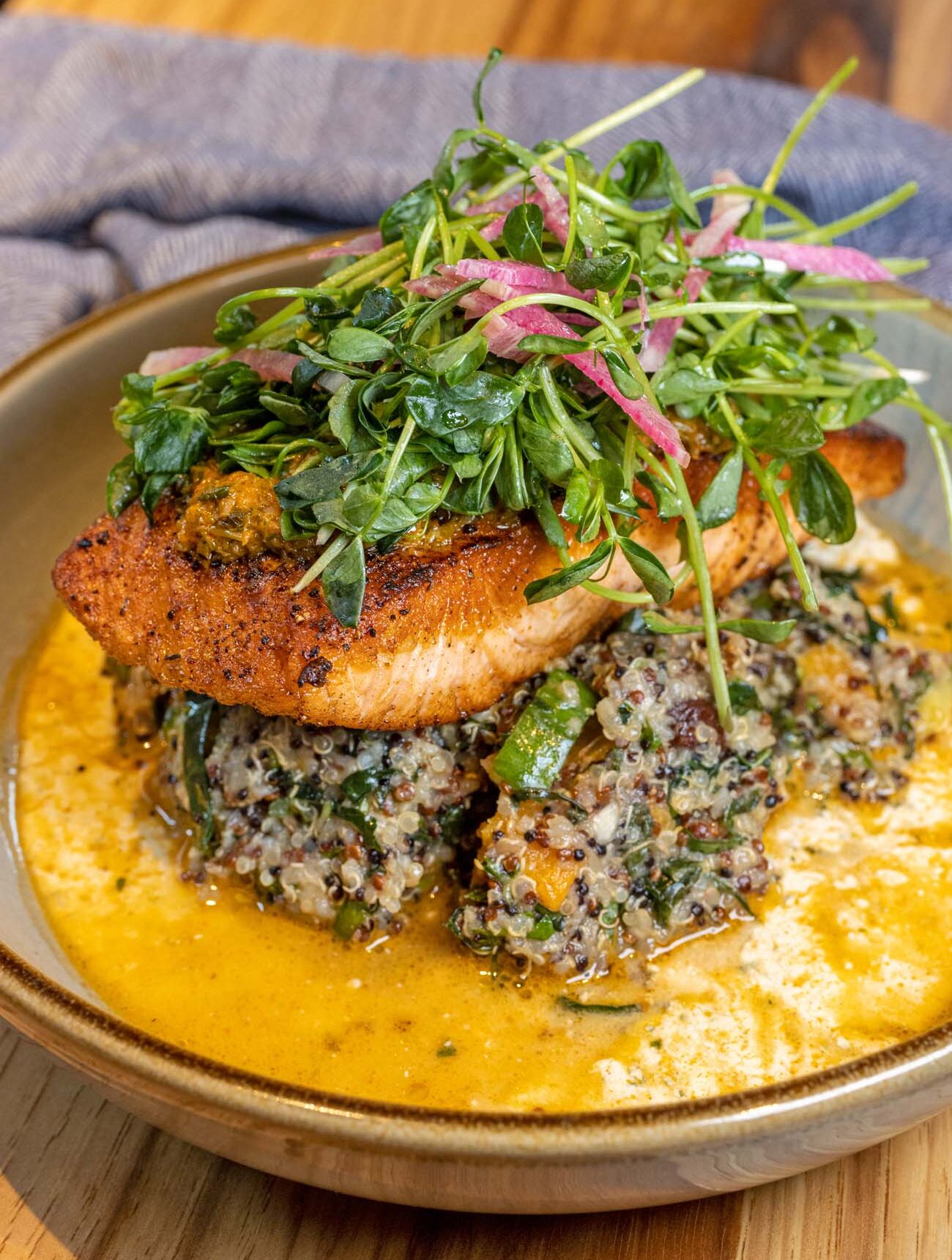 Close-up of an artfully plated salmon dish from Plough Restaurant at the Lancaster Marriott. The perfectly seared salmon fillet with a crispy exterior sits atop a bed of multi-grain salad that appears to include quinoa and fresh herbs. The dish is garnished with a vibrant microgreen and radish salad, and is presented in a shallow bowl with a golden-hued sauce or broth. The elegant presentation on a rustic ceramic plate against a wooden table surface highlights the restaurant's sophisticated farm-to-table concept. This image likely represents the "Roasted Atlantic Salmon" described in the blog post as "the freshest salmon I've had since visiting Canada" with its "perfectly crisp" exterior and "buttery, melt-in-your-mouth interior," accompanied by creative accompaniments that provide "sweet-savory bursts" and showcase the chef's commitment to local sourcing and culinary creativity.