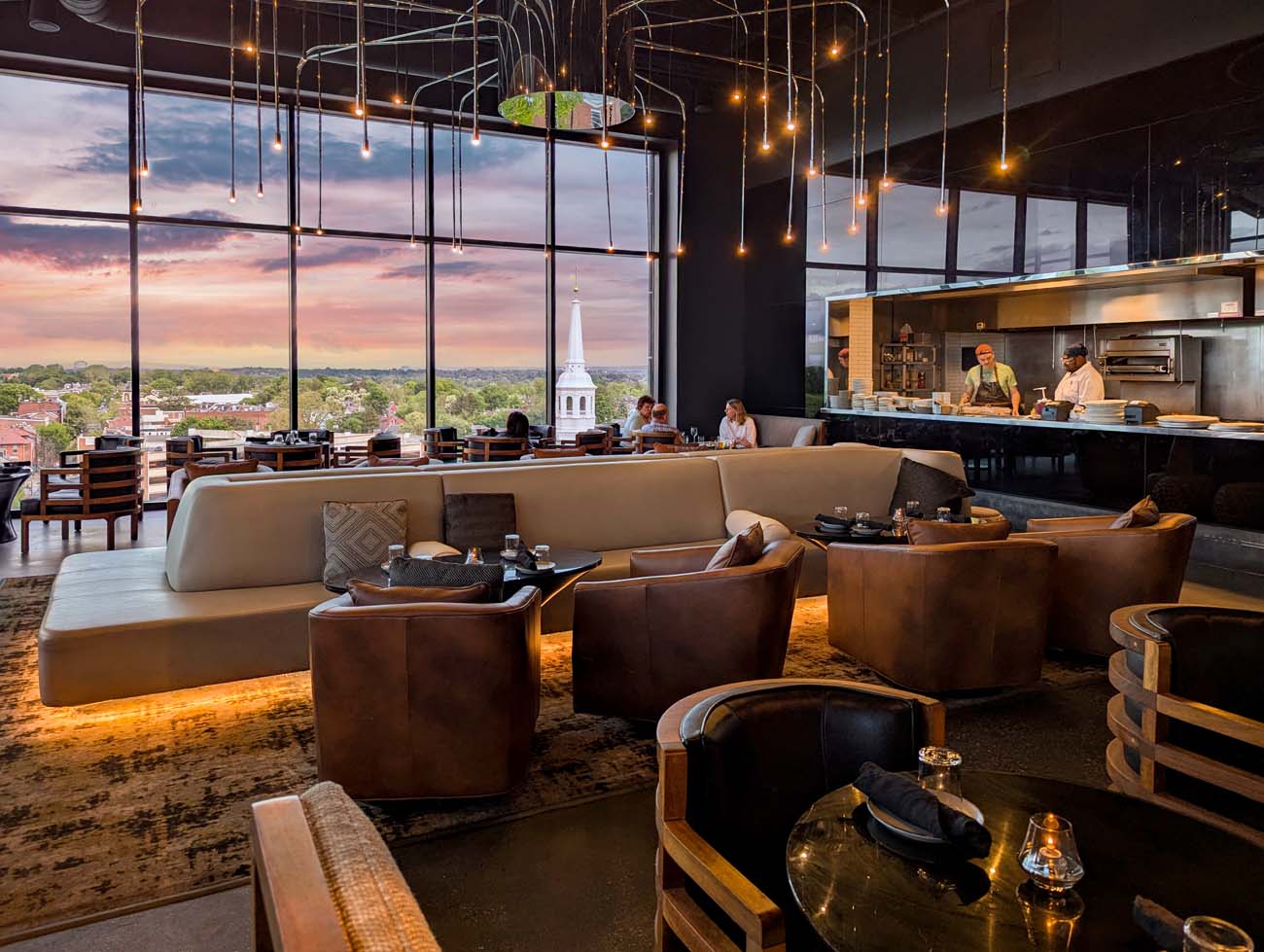 Interior view of The Exchange rooftop restaurant located on the 12th floor of the Lancaster Marriott. The sophisticated space features floor-to-ceiling windows offering panoramic views of downtown Lancaster at sunset, with a church steeple visible against the colorful sky. The restaurant's modern design includes cream-colored curved banquette seating, leather club chairs, and stylish cocktail tables. Dramatic pendant lighting with small Edison bulbs hangs from the ceiling, creating an intimate atmosphere. To the right, an open kitchen where chefs can be seen at work adds to the dynamic dining experience. The space combines upscale design elements with comfortable seating arrangements, embodying what the blog post describes as "that perfect balance between upscale and approachable." This image captures exactly why the author recommended arriving at 5:30pm to "secure a prime spot" for watching the sunset transform the city's historic architecture.