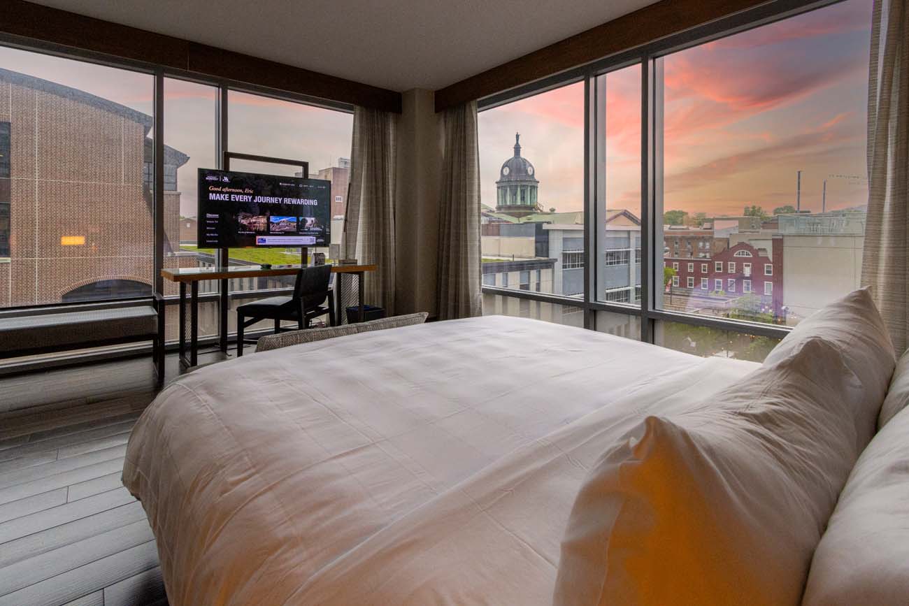 Interior view of a stylish hotel room at the Lancaster Marriott at Penn Square, featuring floor-to-ceiling windows that showcase a stunning sunset view of downtown Lancaster. The room contains a king-sized bed with crisp white linens and multiple pillows, a modern desk with a flatscreen TV, and contemporary furnishings. Through the windows, the iconic domed courthouse or administrative building is visible against the pink and orange sunset sky, demonstrating the hotel's prime location in the heart of downtown Lancaster. The sleek, modern design of the room with its clean lines and neutral palette reflects the "modern luxury" mentioned in the blog post, while the expansive city views illustrate why the author recommends requesting higher floors for the best vistas. This image perfectly captures the room's description of having "floor-to-ceiling windows and sweeping views of the city."Interior view of a stylish hotel room at the Lancaster Marriott at Penn Square, featuring floor-to-ceiling windows that showcase a stunning sunset view of downtown Lancaster. The room contains a king-sized bed with crisp white linens and multiple pillows, a modern desk with a flatscreen TV, and contemporary furnishings. Through the windows, the iconic domed courthouse or administrative building is visible against the pink and orange sunset sky, demonstrating the hotel's prime location in the heart of downtown Lancaster. The sleek, modern design of the room with its clean lines and neutral palette reflects the "modern luxury" mentioned in the blog post, while the expansive city views illustrate why the author recommends requesting higher floors for the best vistas. This image perfectly captures the room's description of having "floor-to-ceiling windows and sweeping views of the city."