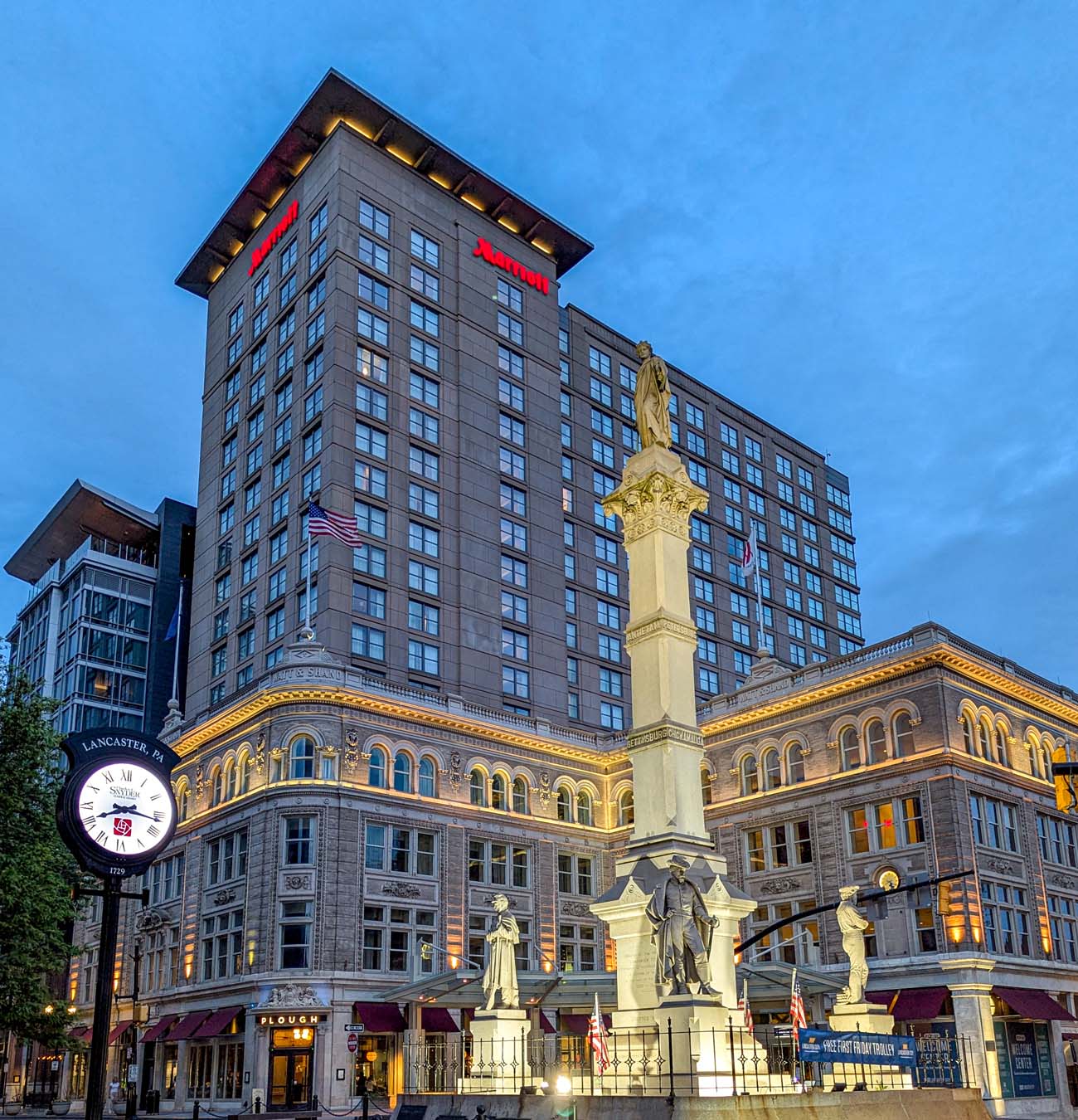 Twilight exterior view of the Lancaster Marriott at Penn Square, capturing the hotel's impressive architectural blend of historic and modern elements. The image shows the beautifully illuminated 1898 Beaux Arts-style Watt & Shand building at the base with its ornate stonework, arched windows, and decorative cornices, seamlessly connected to the modern hotel tower rising above with its contemporary facade and glowing red Marriott signage. In the foreground stands the Soldiers and Sailors Monument, a tall column with statues that commemorates Civil War veterans, dramatically lit against the evening sky. A decorative street clock is visible on the left, and awnings for the Plough restaurant can be seen at street level. This image perfectly illustrates the blog post's description of the hotel as "a seamless blend of the 1898 Beaux Arts-style Watt & Shand building with a modern tower rising behind it," showcasing its prime location at Penn Square in the heart of Lancaster's historic district.