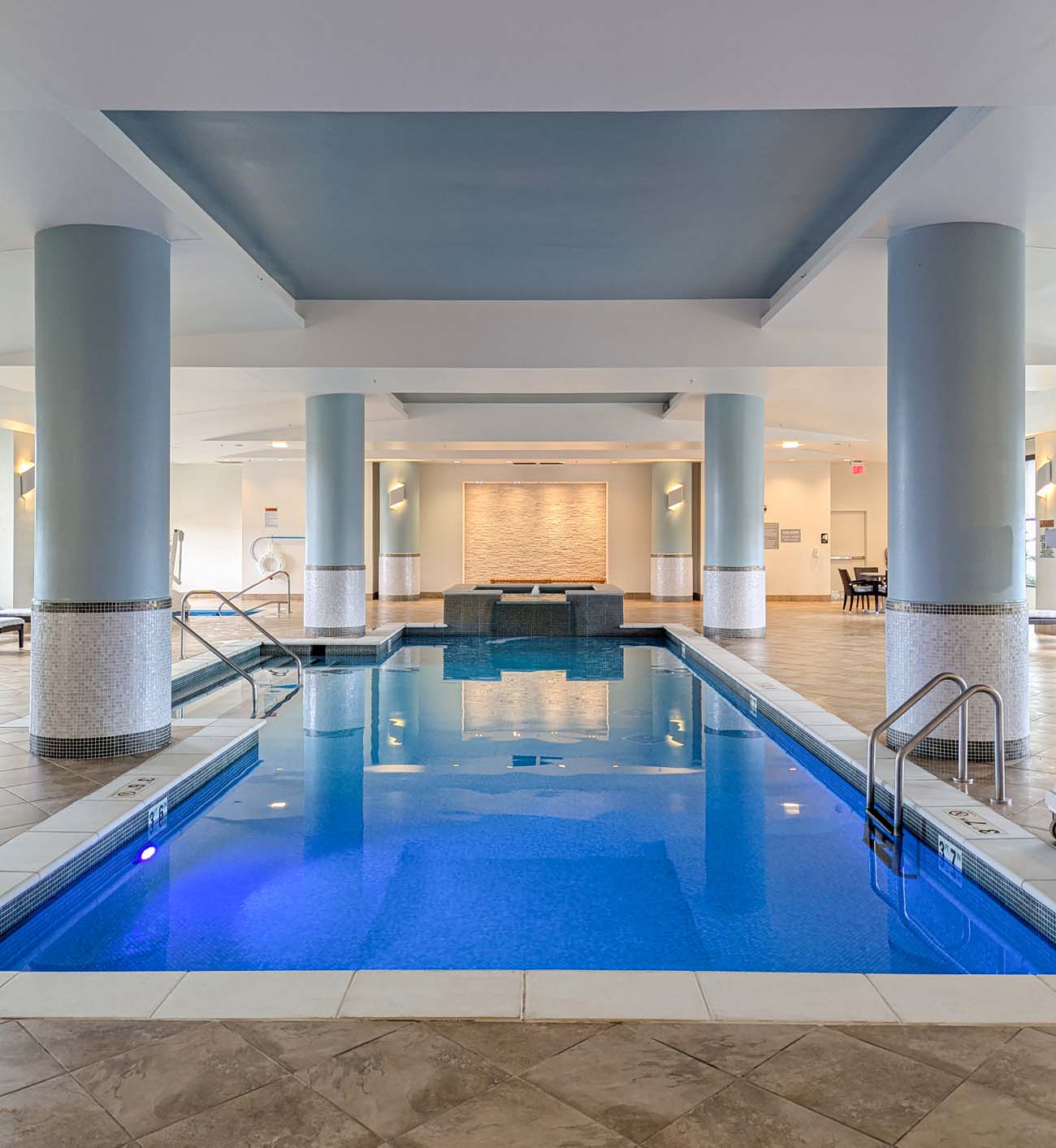 Interior view of the Lancaster Marriott's elegant indoor pool area, featuring a serene rectangular pool with blue-illuminated water surrounded by clean white tile decking. Large pale blue columns with mosaic tile accents support the ceiling, creating a sophisticated atmosphere reminiscent of a luxury resort rather than a typical hotel pool. The space includes built-in steps for pool entry, handrails, and appears to have a hot tub or water feature at the far end. The minimalist design with its cool color palette of whites and blues creates a tranquil oasis within the hotel. This image illustrates what the blog post describes as the "resort-like atmosphere" of the hotel's pool area that "surprised" the author with its upscale ambiance and thoughtful design elements, making it an unexpected amenity in a city hotel.