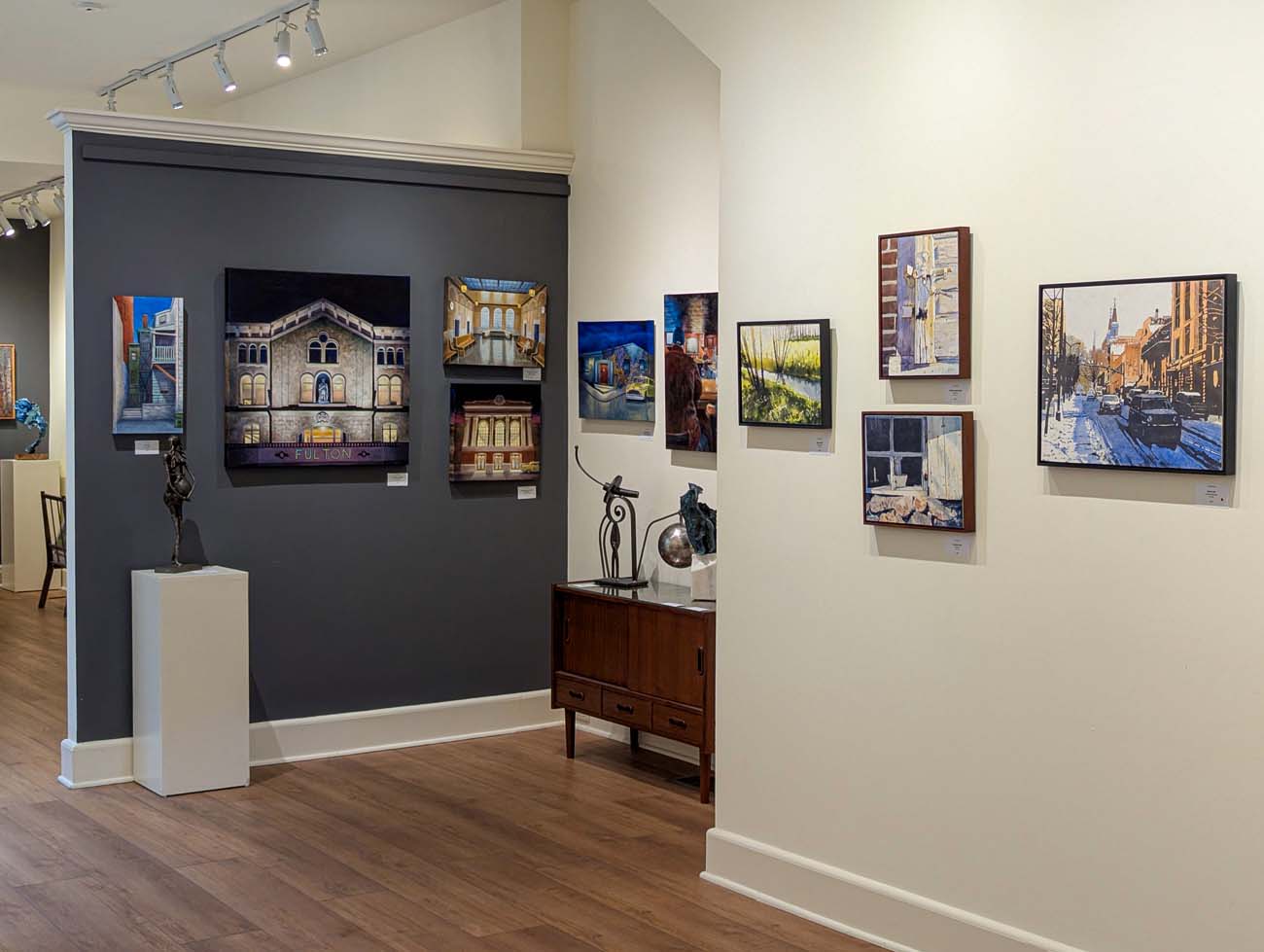 Interior view of Red Raven in downtown Lancaster's Gallery Row district with professionally displayed artwork on light and dark accent walls. The gallery features paintings of Lancaster landmarks, including a prominent painting of the historic Fulton Theatre facade in the center of the dark gray feature wall. Multiple cityscape paintings showcase Lancaster's architectural heritage and street scenes. Small sculptures are displayed on pedestals, and the space has polished wood floors with track lighting that highlights the artwork. The gallery exemplifies the curated art experience available along North Prince Street, where visitors can view and purchase work from local and regional artists.