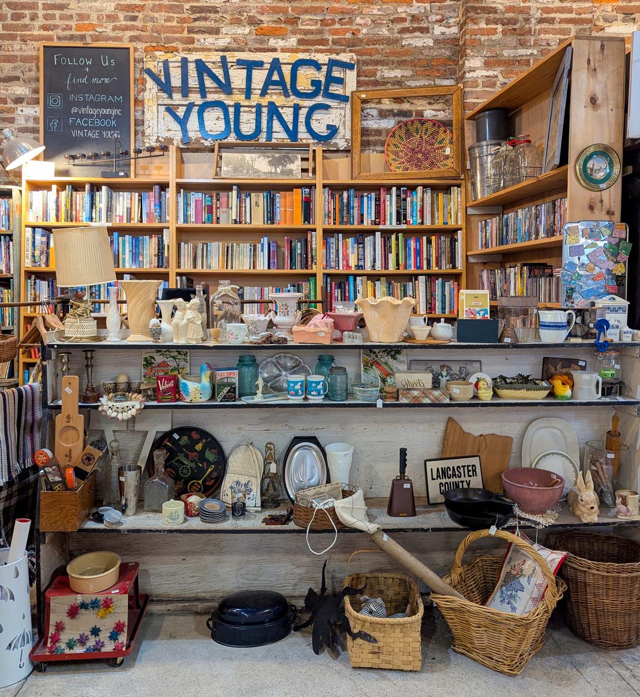 Interior of the Vintage Young shop in downtown Lancaster featuring floor-to-ceiling bookshelves and displays of antiques and collectibles. The shop has an exposed brick wall with a prominent "VINTAGE YOUNG" blue sign. Multiple shelves are filled with books, while display counters showcase an eclectic collection of vintage items including ceramics, dishes, glassware, baskets, and decorative pieces. A chalkboard sign invites customers to follow the shop on social media. The space embodies the charm of Lancaster's independent retailers, offering a nostalgic shopping experience where visitors can hunt for unique treasures among carefully curated vintage goods and literature.