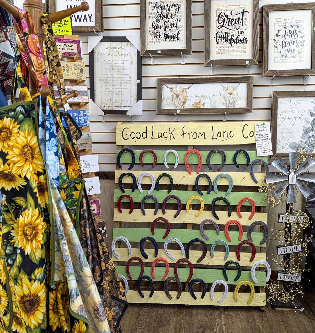 Interior display at an Amish Country gift shop featuring a mix of faith-themed artwork and local crafts. The wall is lined with framed prints featuring inspirational sayings and hymn lyrics including "Amazing Grace" and "Great Faithfulness" in elegant calligraphy. The centerpiece is a colorful display of painted horseshoes arranged on a wooden board labeled "Good Luck from Lanc. Co." with horseshoes in various colors including red, blue, green, and black. To the left, sunflower-patterned fabric is visible, and decorative signs reading "Faith," "Hope," and "Love" hang nearby. This display represents the blend of religious themes and rural traditions often found in Lancaster County's gift shops that appeal to tourists seeking authentic souvenirs