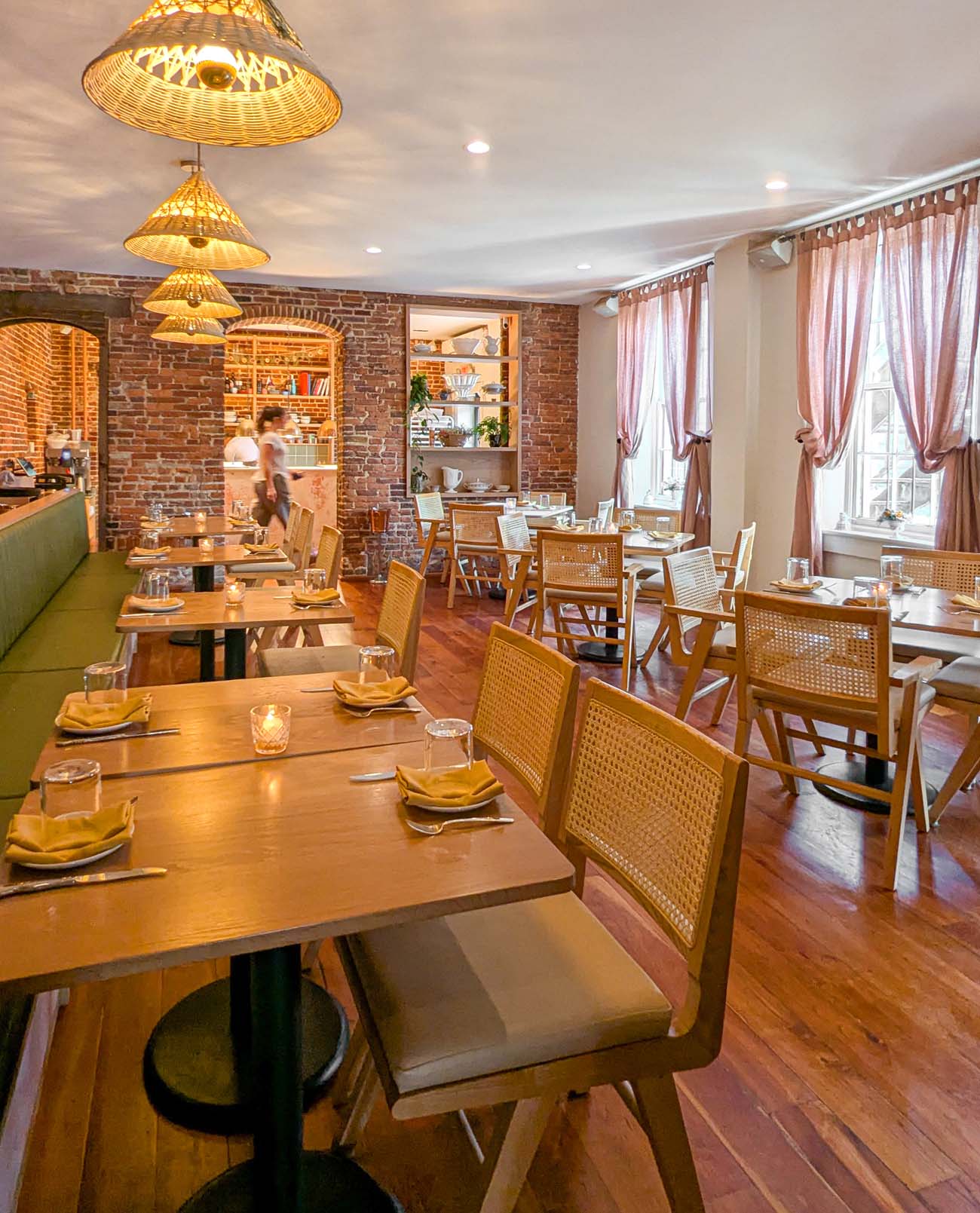 The elegant dining room of Passerine restaurant in downtown Lancaster, featuring exposed brick walls, warm wooden floors, and natural cane-backed chairs at wooden tables. Woven pendant lights hang from the ceiling, casting a warm glow over the space. The left side features a green banquette seating area, while tables are carefully set with folded yellow napkins, glassware, and candles. Pink curtains soften the natural light coming through windows on the right side. A server can be seen moving through the space, suggesting the attentive service. This New York Times-recognized restaurant embodies Lancaster's sophisticated culinary scene, with its thoughtful design balancing historic architectural elements with contemporary dining comfort.
