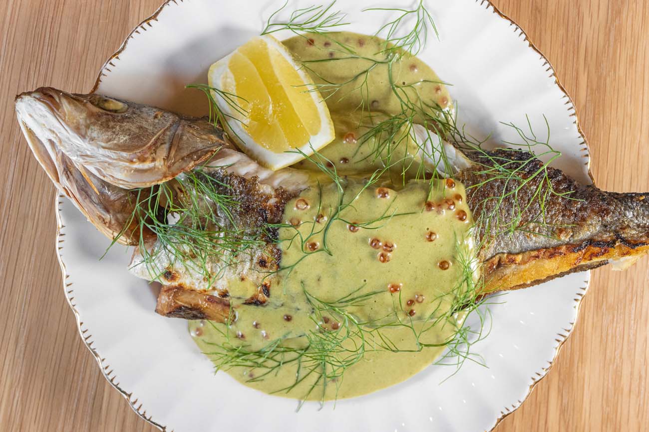 Beautifully presented whole branzino on a white scalloped-edge plate at Passerine restaurant. The fish is served with its head and tail intact, covered in delicate fresh dill and a light green sauce dotted with mustard seeds. A fresh lemon wedge garnishes the fish, adding brightness to the dish. The presentation exemplifies Passerine's seasonal, locally-sourced culinary philosophy that earned them recognition from The New York Times. The elegant plating on the simple white dish against a wooden table surface highlights the restaurant's approach of letting high-quality ingredients speak for themselves, demonstrating the sophisticated dining experiences available in downtown Lancaster.