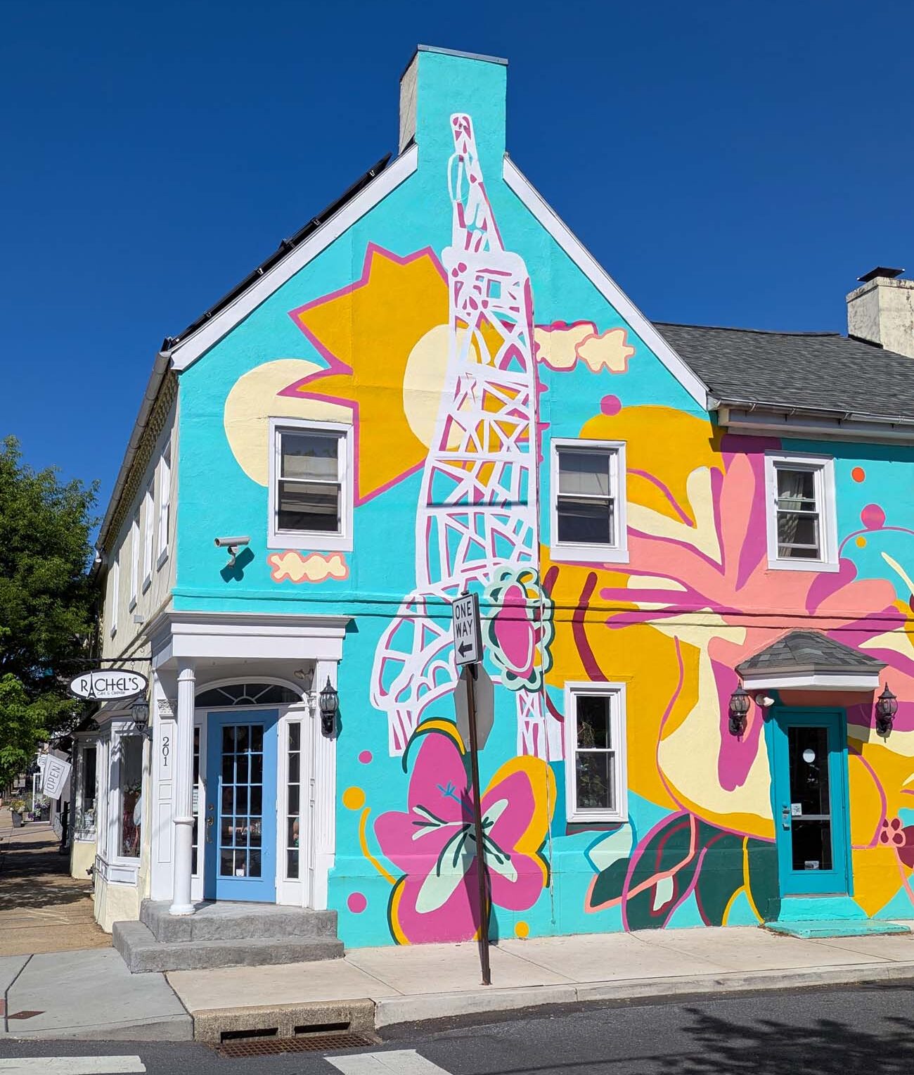 A striking turquoise building housing Rachel's Café & Creperie, adorned with a bold, colorful mural featuring oversized pink and yellow flowers, abstract shapes, and a white Eiffel Tower-like structure. The corner building has white trim, multiple windows, and a blue entrance door. "Rachel's" signage is visible on the left side of the building. The vibrant artwork transforms this historic structure into a landmark that stands out against the clear blue sky, exemplifying Lancaster's blend of historic architecture and contemporary artistic expression. Located on West Walnut Street, this colorful café is known for its massive handheld crepes and has become a popular breakfast and brunch destination in downtown Lancaster.