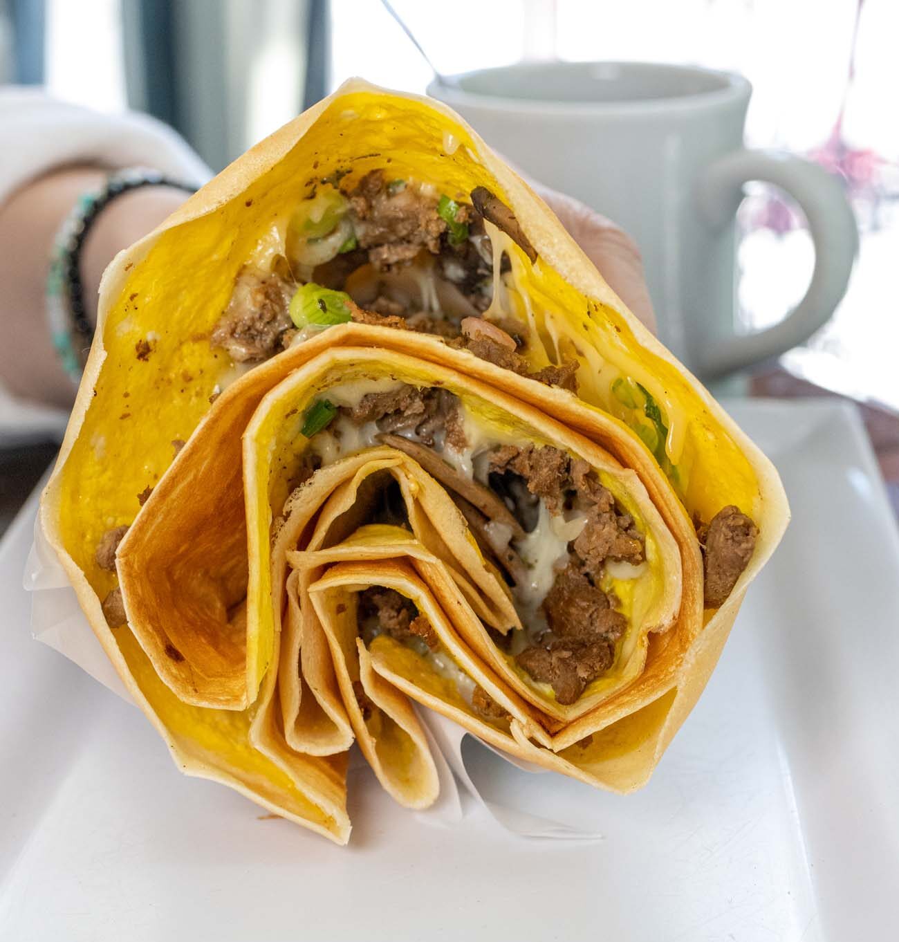 Close-up of a large savory crepe from Rachel's Café & Creperie in downtown Lancaster, shown cut in half to reveal its generous filling of seasoned ground meat, melted cheese, and green onions. The golden-brown crepe is folded into a distinctive multilayered pattern and served on a white square plate. In the background, a coffee mug is visible, suggesting the typical breakfast or brunch setting. This substantial crepe represents the café's signature "hoagie-sized" handheld meals that have made Rachel's a popular dining destination for 18 years. The crepe showcases the café's unique approach to French-inspired cuisine adapted to hearty American appetites, demonstrating Lancaster's diverse culinary scene.