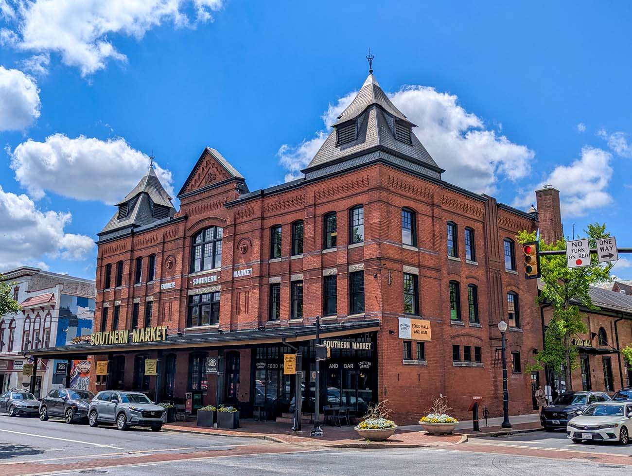 The impressive red brick exterior of Southern Market, a historic building in downtown Lancaster. Built in 1888, the structure features distinctive Victorian architecture with ornate details, including two decorative towers with slate roofs. The building has been renovated into a modern food hall while preserving its historic character. Large windows line the façade, and the name "SOUTHERN MARKET" is prominently displayed across the front. The street-level entrance features "FOOD HALL AND BAR" signage. Located just a block from the Lancaster Marriott, this repurposed building houses over a dozen chef stations offering global cuisines from Moroccan to Middle Eastern, Latin, and more. The bright blue sky with puffy white clouds accentuates the building's rich red brick exterior.