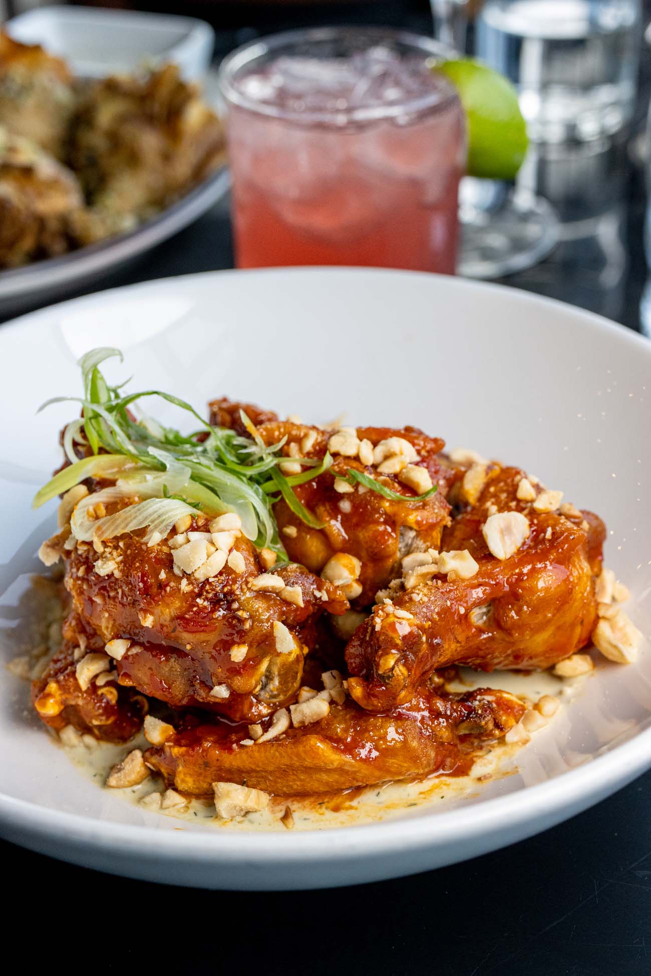 A close-up photograph of glazed chicken wings served in a white bowl. The wings are coated in a rich, glossy reddish-brown sauce that appears to be the Calabrian chili glaze mentioned in the blog post about The Exchange rooftop restaurant in Lancaster. The dish is topped with chopped toasted hazelnuts and thinly sliced green onions, adding texture and a fresh garnish. In the background, slightly out of focus, there's a vibrant red cocktail with ice and what appears to be a lime garnish, possibly the Mezcolada cocktail also mentioned in the blog. This signature appetizer from The Exchange rooftop bar at the Lancaster Marriott features an addictive balance of sweetness, heat, and crunch from the hazelnuts that made it a standout dish during the author's visit.