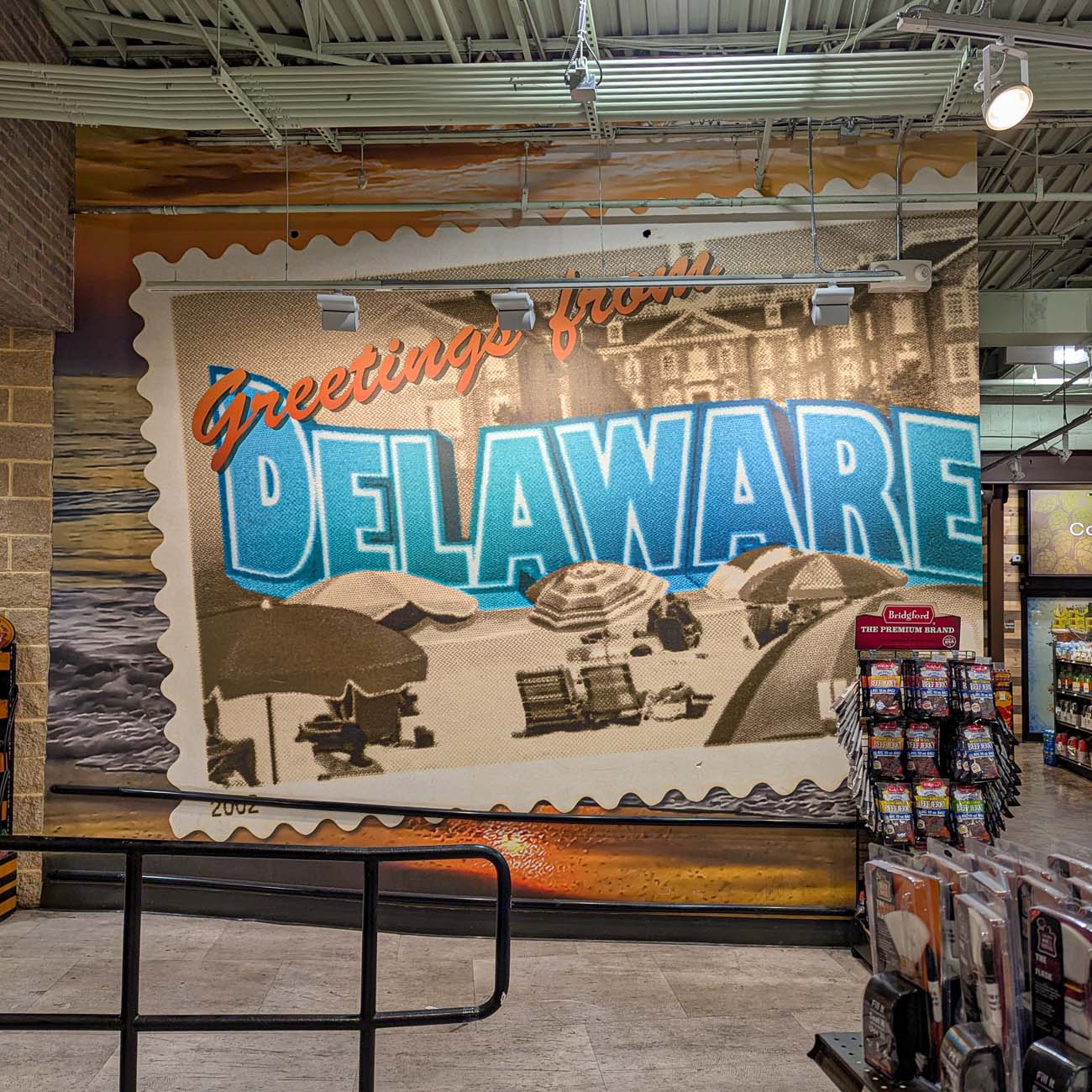 Interior of a Total Wine store featuring a large decorative wall mural designed like a vintage postcard that reads "Greetings from DELAWARE" in bright orange and blue lettering, encouraging to shop for Delaware craft beverages. The postcard design shows a beach scene with umbrellas and beach chairs. The mural is mounted on a brick wall within the retail space with merchandise displays visible in the foreground.