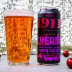1911 Cherry Hard Cider poured and ready to enjoy. A tall glass filled with golden amber hard cider sits next to a black can of 1911 Established "Light Up The Night" Cherry Premium Small Batch Hard Cider by Beak & Skiff. The can features bold pink and purple lettering on a black background. Several fresh red cherries are scattered around the base of the glass and can on a white surface, with a sparkling bokeh background creating an elegant atmosphere.
