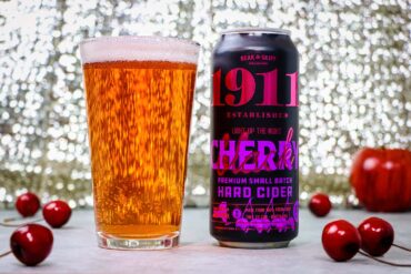 1911 Cherry Hard Cider poured and ready to enjoy. A tall glass filled with golden amber hard cider sits next to a black can of 1911 Established "Light Up The Night" Cherry Premium Small Batch Hard Cider by Beak & Skiff. The can features bold pink and purple lettering on a black background. Several fresh red cherries are scattered around the base of the glass and can on a white surface, with a sparkling bokeh background creating an elegant atmosphere.
