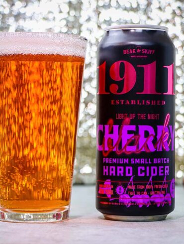 1911 Cherry Hard Cider poured and ready to enjoy. A tall glass filled with golden amber hard cider sits next to a black can of 1911 Established "Light Up The Night" Cherry Premium Small Batch Hard Cider by Beak & Skiff. The can features bold pink and purple lettering on a black background. Several fresh red cherries are scattered around the base of the glass and can on a white surface, with a sparkling bokeh background creating an elegant atmosphere.