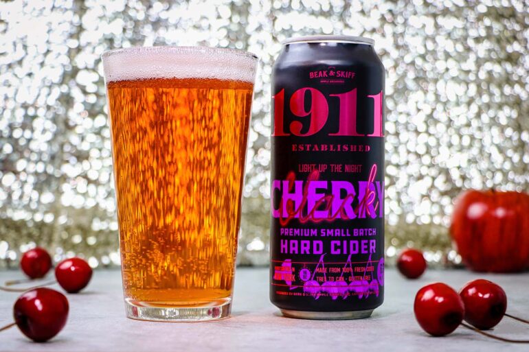 1911 Cherry Hard Cider poured and ready to enjoy. A tall glass filled with golden amber hard cider sits next to a black can of 1911 Established "Light Up The Night" Cherry Premium Small Batch Hard Cider by Beak & Skiff. The can features bold pink and purple lettering on a black background. Several fresh red cherries are scattered around the base of the glass and can on a white surface, with a sparkling bokeh background creating an elegant atmosphere.