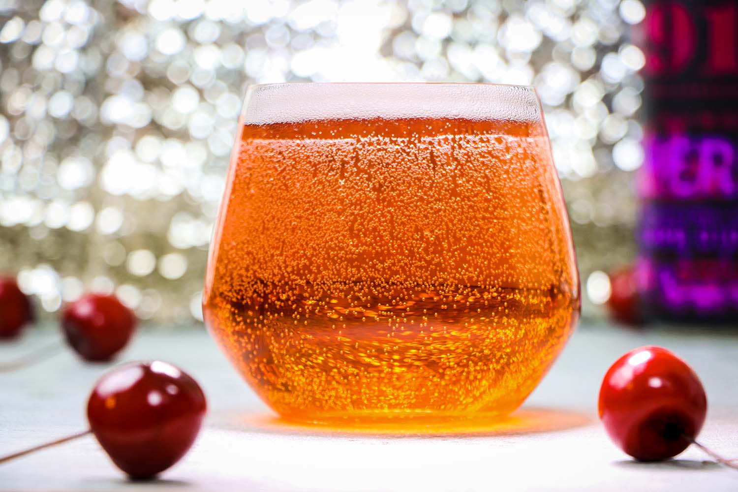 A close-up shot of golden amber hard cider in a stemless wine glass, showcasing active carbonation with thousands of tiny bubbles rising through the liquid. The cider has a beautiful gradient from deep amber at the bottom to lighter golden tones at the top with a thin layer of foam. Red cherries are visible in the foreground and background, with soft bokeh lighting effects creating a warm, inviting ambiance.