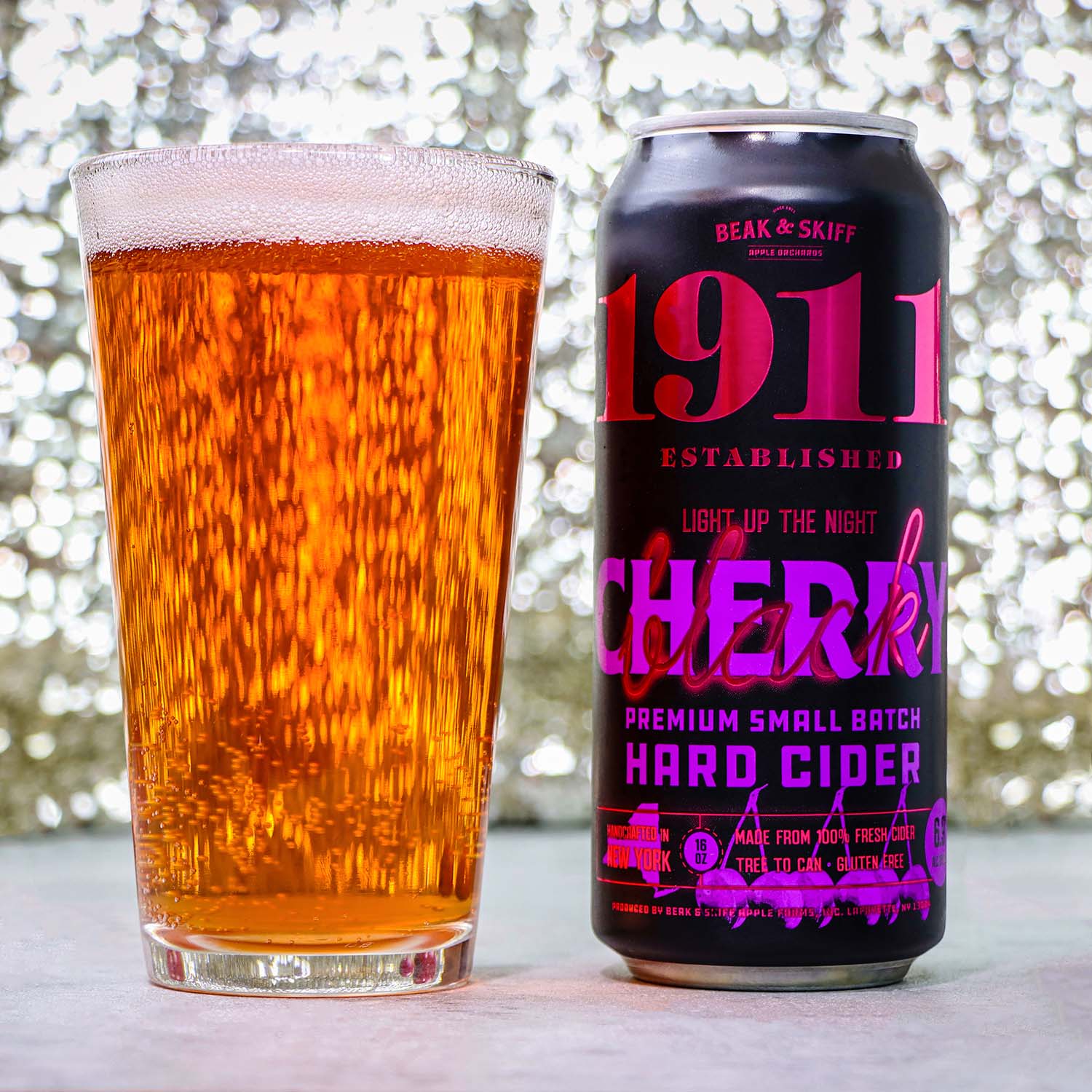 A pint glass filled with golden amber hard cider displaying vigorous carbonation with streams of bubbles rising throughout the liquid, topped with a thin white foam head, positioned next to a black can of 1911 Established "Light Up The Night" Black Cherry Premium Small Batch Hard Cider by Beak & Skiff. The can features bold pink and purple gradient lettering on a matte black background with product details including "Made from 100% Fresh Cold Pressed New York Apples" and "Gluten Free." Both items sit on a light wooden surface against a dreamy bokeh background of soft, blurred lights creating an atmospheric and premium presentation.