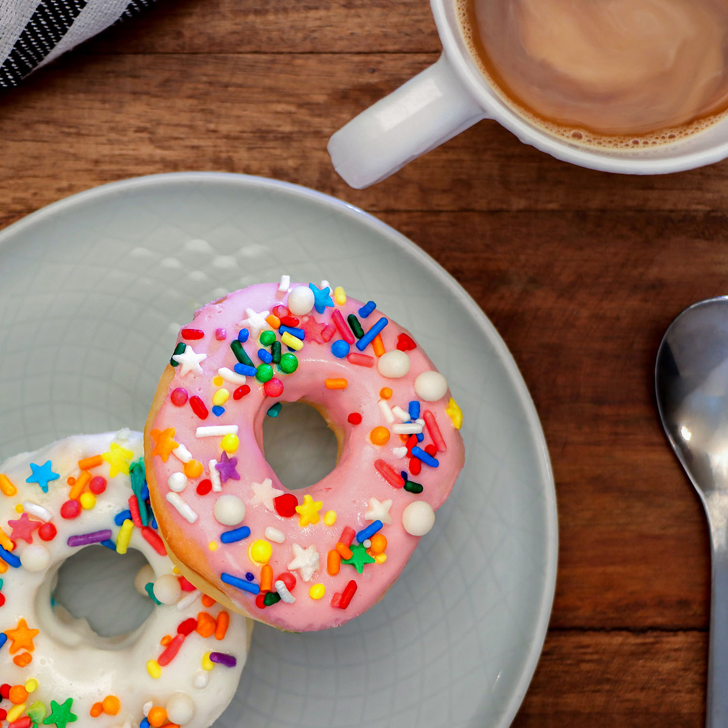 Two glazed air fried donuts with rainbow sprinkles on a light blue ceramic plate next to a white coffee mug filled with latte, photographed from above on a wooden table surface.