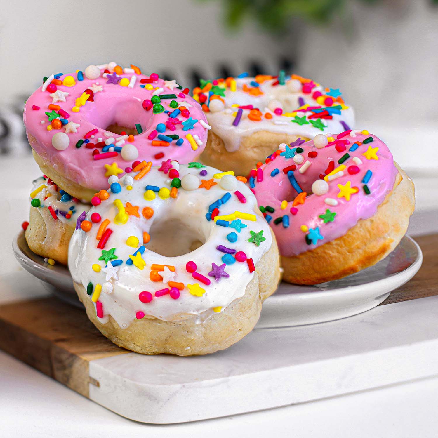 Close-up view of glazed air fryer donuts with pink and white icing topped with colorful rainbow sprinkles including jimmies, nonpareils, and star-shaped decorations, displayed on a white plate.
