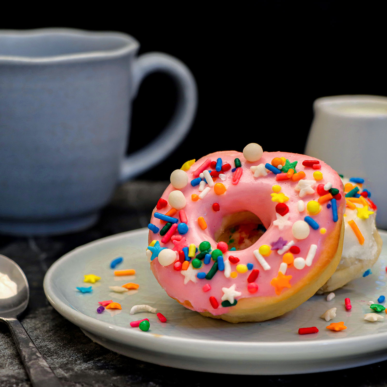 Single pink-glazed air fryer donut with multicolored sprinkles on a white plate with scattered sprinkles around it, alongside a blue coffee mug in soft focus background, showing the donut's air fried fluffy texture.
