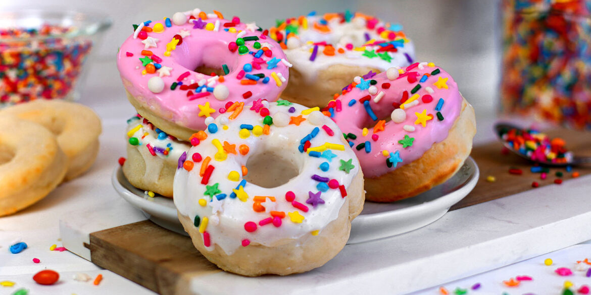 Multiple air fryer donuts with pink and white glazes covered in rainbow sprinkles arranged on a white marble cutting board, with jars of colorful sprinkles visible in the background and plain unglazed donuts to the side.