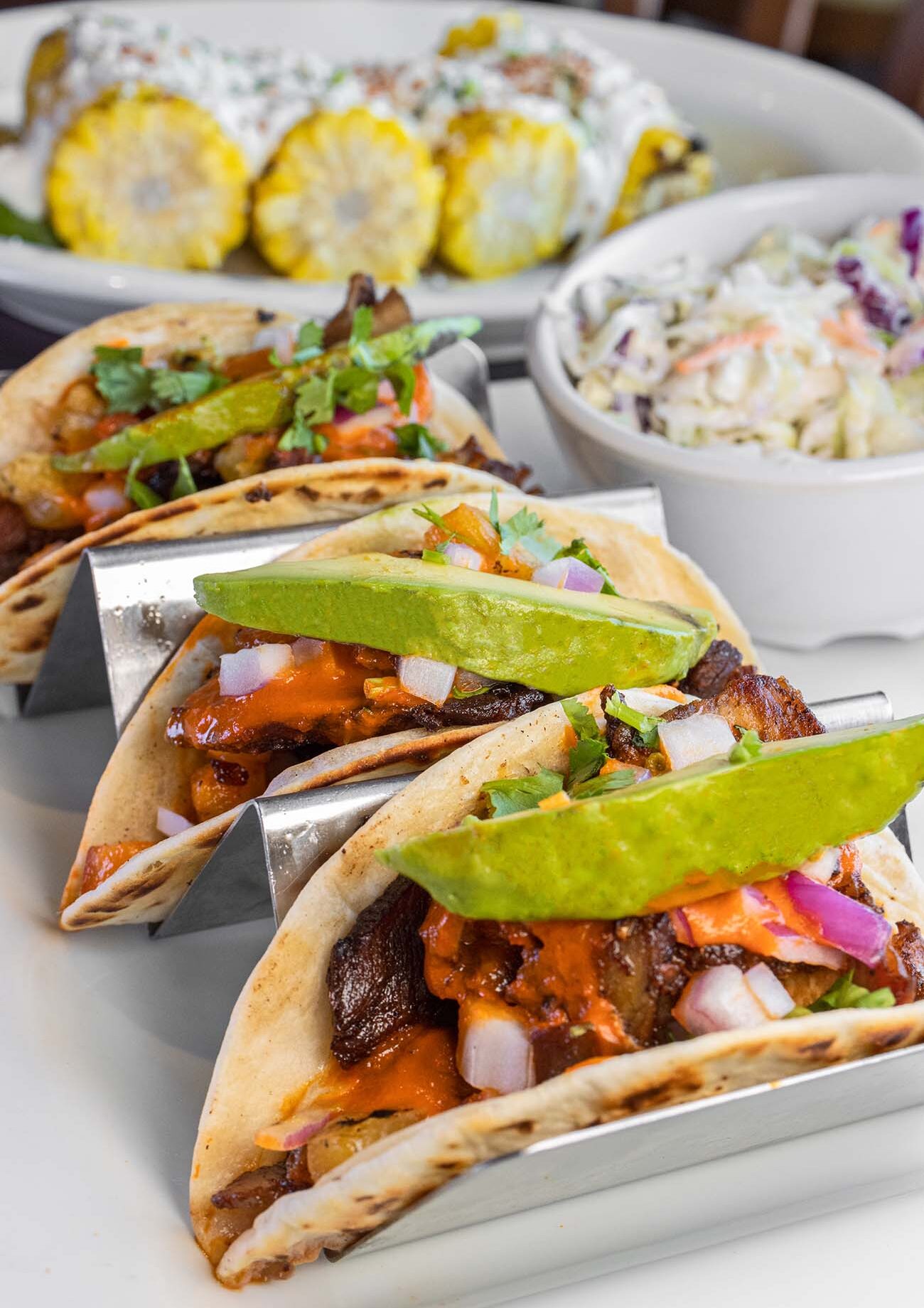 Three soft flour tortillas filled with al pastor pork, topped with fresh avocado slices, diced red onions, cilantro, and orange sauce, served in a metal taco holder alongside grilled corn on the cob and coleslaw at Buttonwood Grill in Peddler's Village