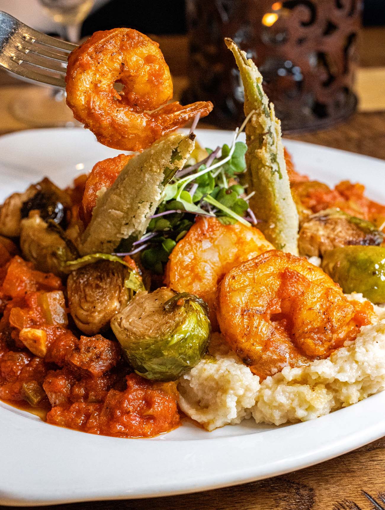 Shrimp and Grits with fried okra towers on a rustic wooden table. Warm glow.. One shrimp on a fork. Candle in the background.