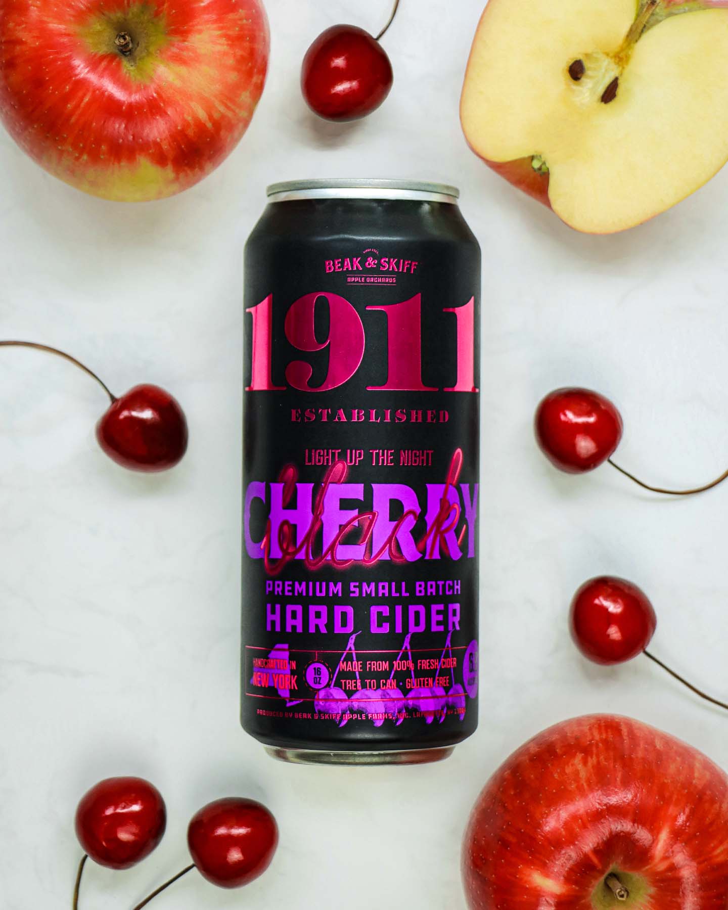 An overhead flat lay composition featuring a black can of 1911 Black Cherry Hard Cider as the centerpiece, surrounded by fresh red apples (one whole, one cut in half showing the yellow flesh), multiple fresh red cherries with stems, all arranged on a clean white background. The can's vibrant pink and purple branding stands out against the natural red tones of the fruit, creating an appealing product shot that highlights the cider's fruit-forward nature.