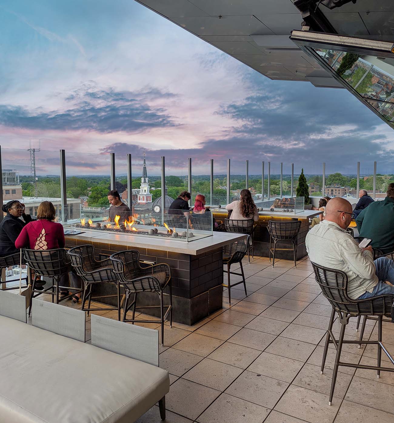 The Exchange Lancaster's outdoor rooftop terrace featuring modern fire pit tables with glass wind barriers, black wire chairs, and panoramic views of downtown Lancaster's church spires and historic buildings during a pink and blue sunset sky