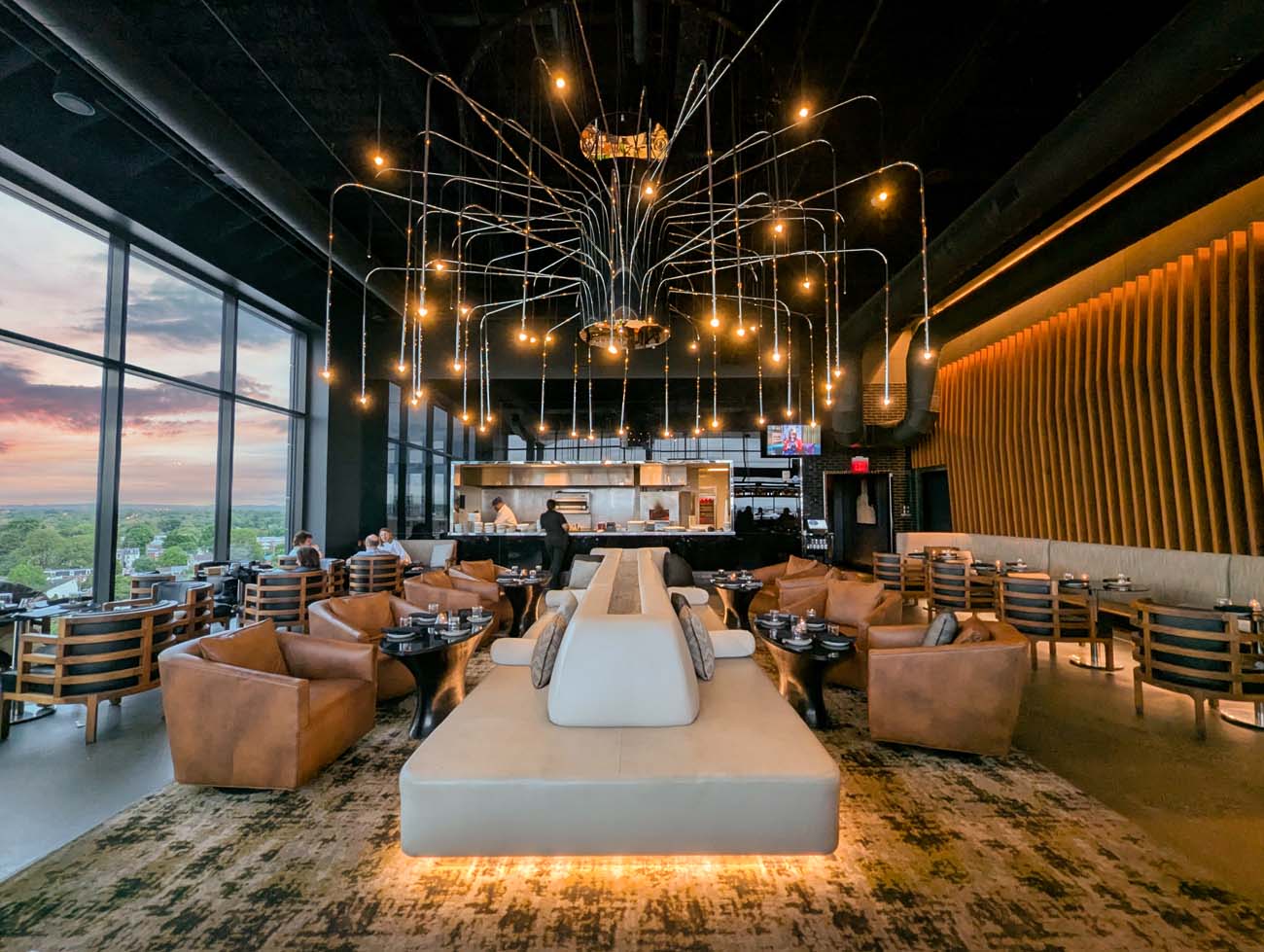 Interior of The Exchange Lancaster showing the modern dining room with dramatic cascading light fixture chandelier, tan leather seating, open kitchen with chefs at work, floor-to-ceiling windows overlooking Lancaster, and warm sunset lighting.