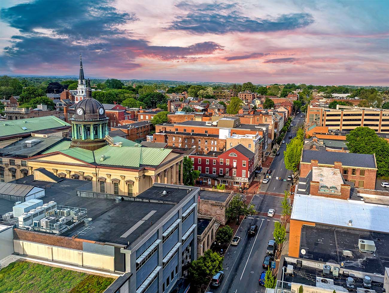 Aerial view from The Exchange Lancaster showing downtown Lancaster's historic architecture including the green-domed courthouse, Trinity Lutheran's white spire, brick buildings, tree-lined streets, and rolling farmland extending to the horizon under a dramatic pink and blue sunset sky.