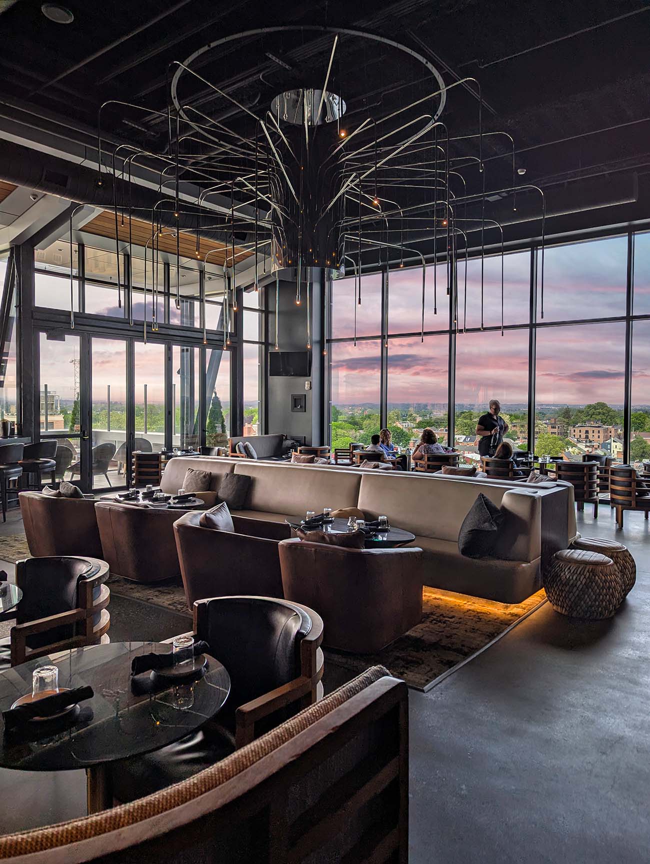 The Exchange Lancaster's interior seating area with curved tan leather banquettes, round cocktail tables, dramatic pendant lighting, and expansive floor-to-ceiling windows showcasing views of Lancaster's church spires and countryside during sunset.