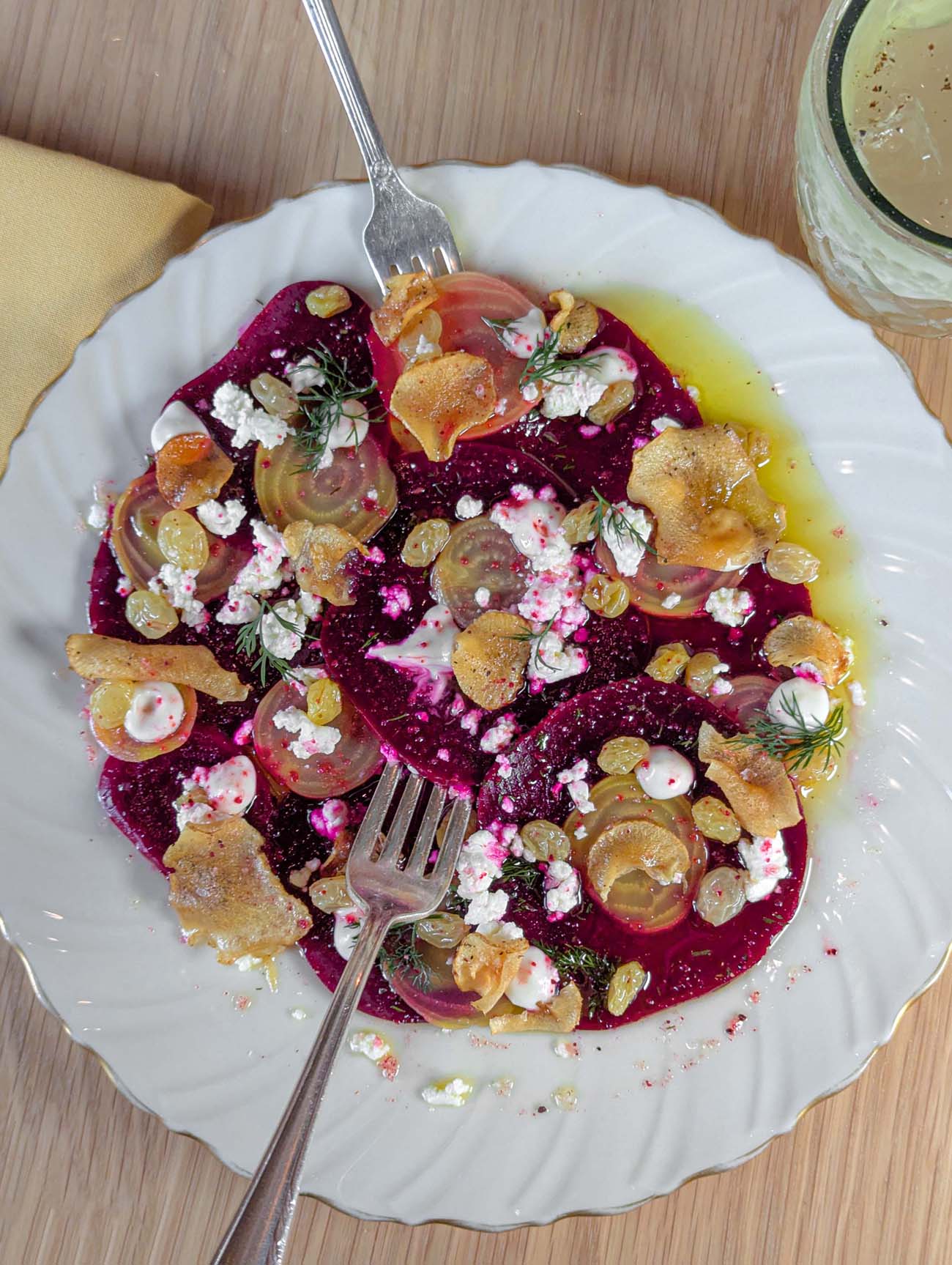 A white scalloped plate containing vibrant beetroot carpaccio with thin slices of deep purple and golden beets arranged artfully, topped with crumbled white goat cheese, golden pickled sultanas, crispy sunchoke chips, fresh dill garnish, and a drizzle of olive oil on a wooden table at Passerine Lancaster