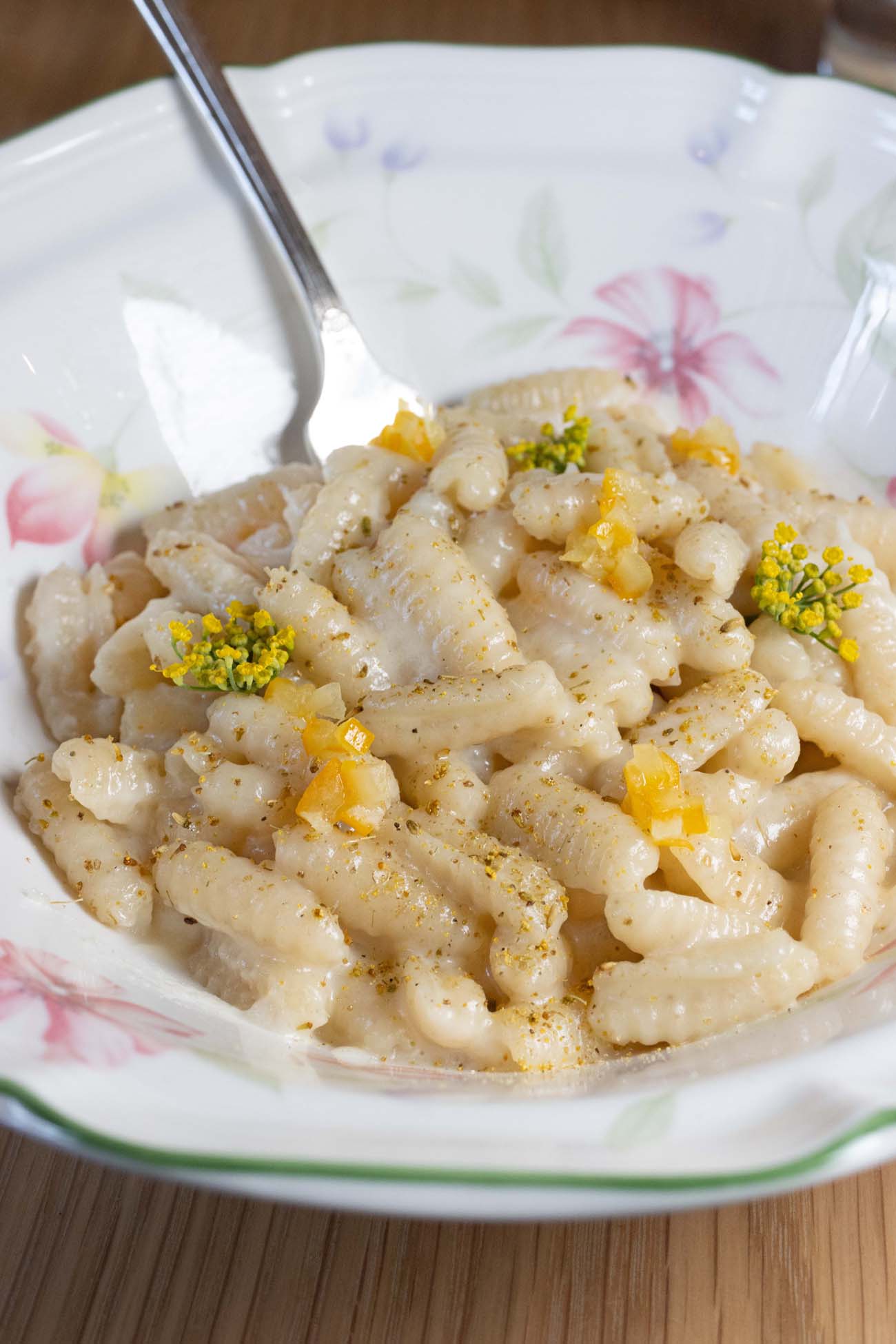 A floral-patterned china bowl filled with handmade gnocchi sardi pasta tossed in pecorino cheese with bright yellow preserved lemon pieces, small yellow fennel flowers as garnish, and freshly cracked black pepper, served on a wooden table