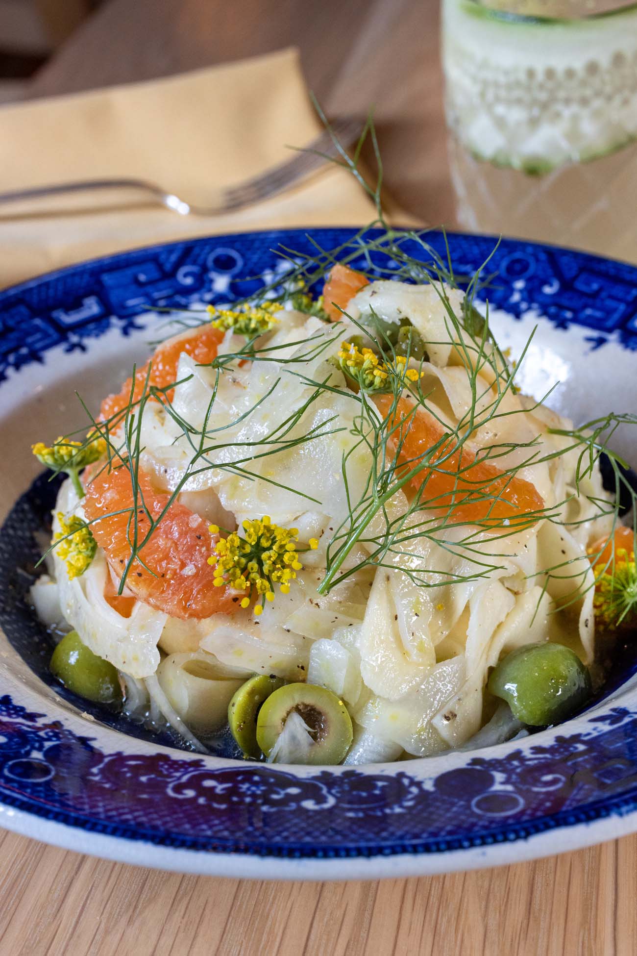 A blue and white patterned ceramic bowl containing ribbons of white parsnip mixed with bright orange citrus segments, green castelvetrano olives, fresh fennel fronds, yellow fennel flowers, and a drizzle of golden smoked oil