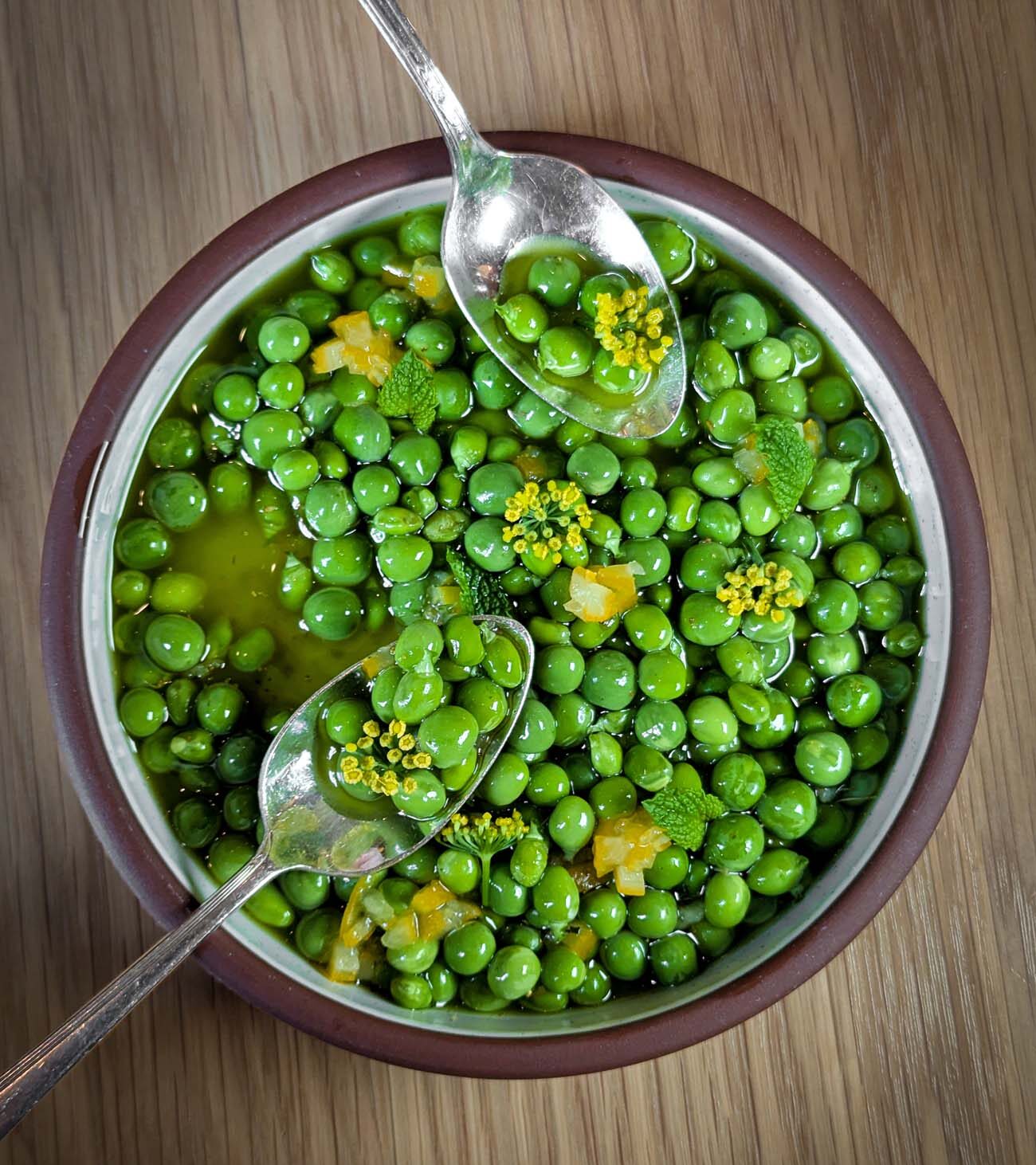 A brown ceramic bowl filled with bright green fresh peas swimming in an emerald green broth, garnished with yellow fennel pollen, fresh mint leaves, diced preserved lemon, and served with two silver spoons for sharing at Passerine Lancaster