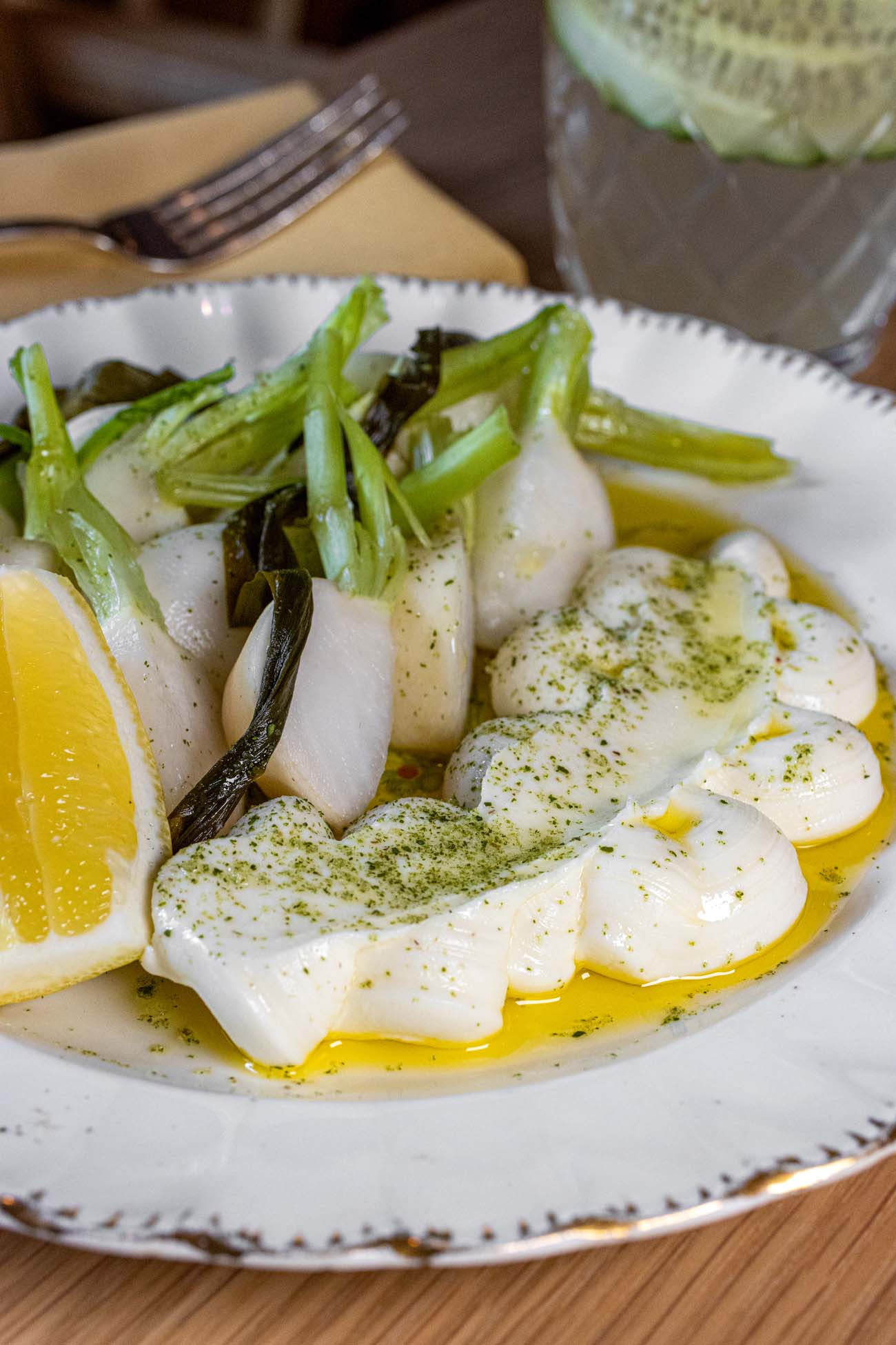 A white scalloped-edge plate containing sliced white Hakurei turnips topped with dollops of whipped ricotta dusted with green herb powder, accompanied by sautéed spring greens and ramps, drizzled with golden olive oil, and garnished with fresh orange segments at Passerine Lancaster