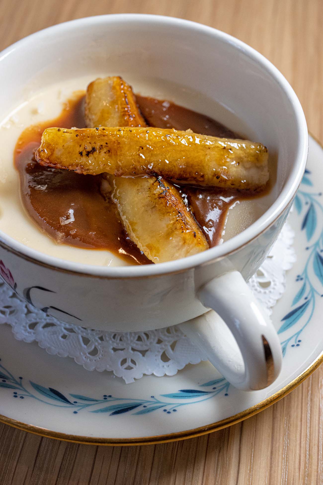 A white ceramic teacup with blue floral pattern and gold rim containing white chocolate panna cotta topped with caramelized banana pieces in rich amber caramel sauce, served on a matching saucer with decorative lace doily at Passerine Lancaster, showcasing their refined dessert presentation