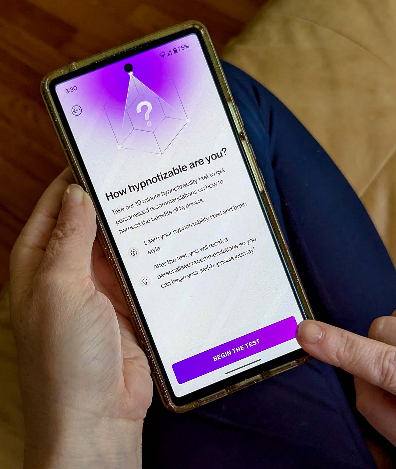 Photo of hands holding a smartphone displaying the Reveri app's "How hypnotizable are you?" assessment screen. The phone shows a purple gradient background with a geometric crystal-like icon containing a question mark. Text explains "Take our 10 minute hypnotizability test to get personalized recommendations on how to harness the benefits of hypnosis." Additional text mentions learning your hypnotizability level and brain style, with personalized recommendations to follow. A purple "BEGIN THE TEST" button is visible at the bottom. The phone appears to be held over someone's lap wearing dark clothing