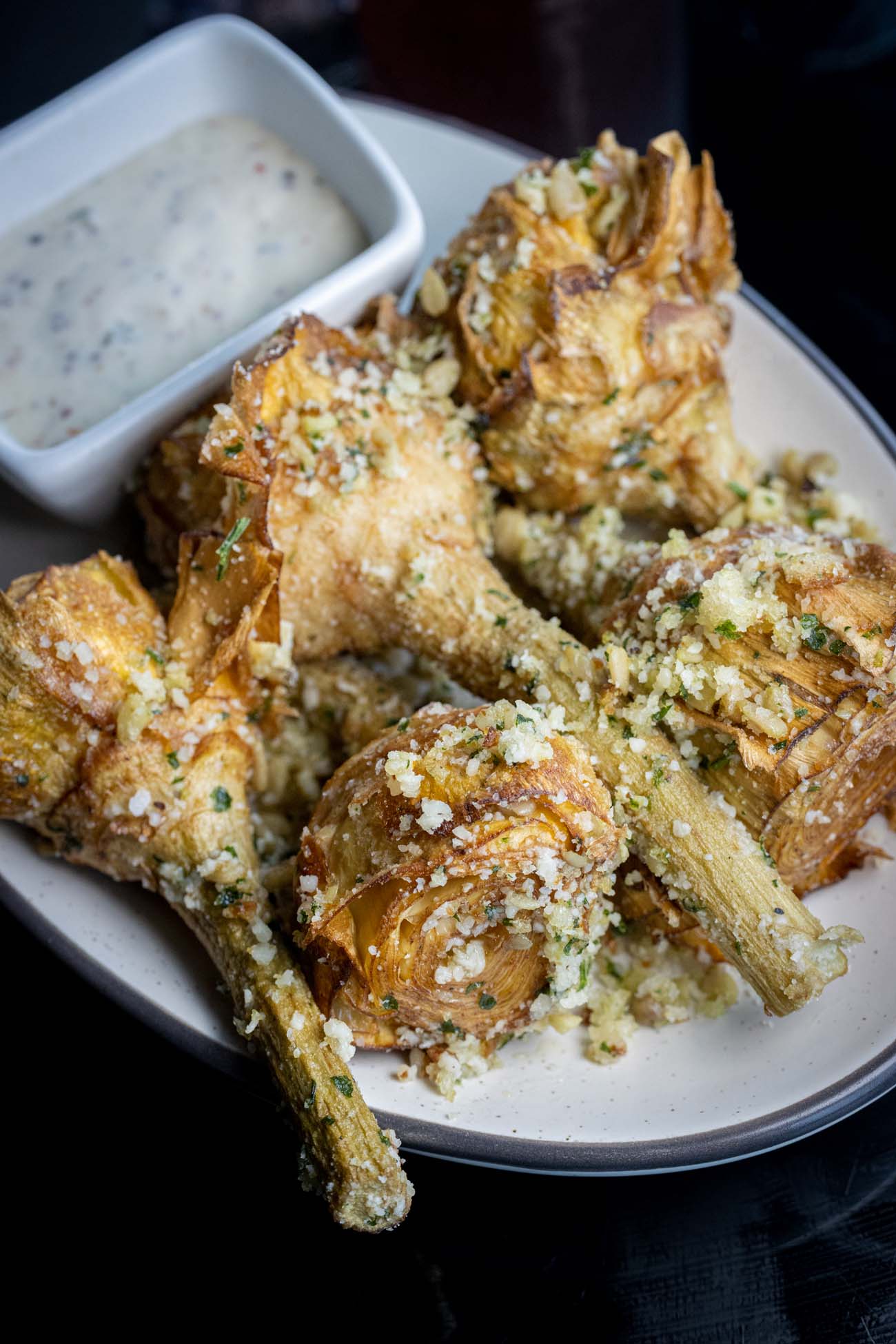 Crispy Baby Artichokes from The Exchange Lancaster served on a white plate, featuring golden-brown fried artichoke halves dusted with grated parmesan cheese and fresh herbs, accompanied by a small bowl of bright citrus aioli dipping sauce.