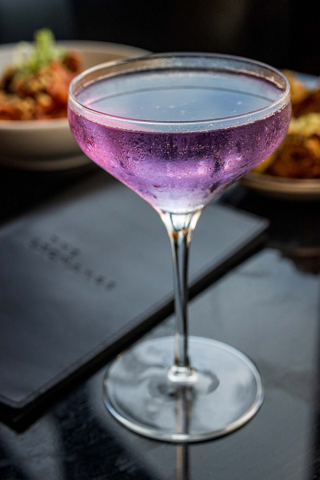 Close-up of a vibrant purple cocktail from The Exchange Lancaster served in a coupe glass with a sugared rim and bubbles throughout the drink, sitting on a dark table with blurred food plates in the background.